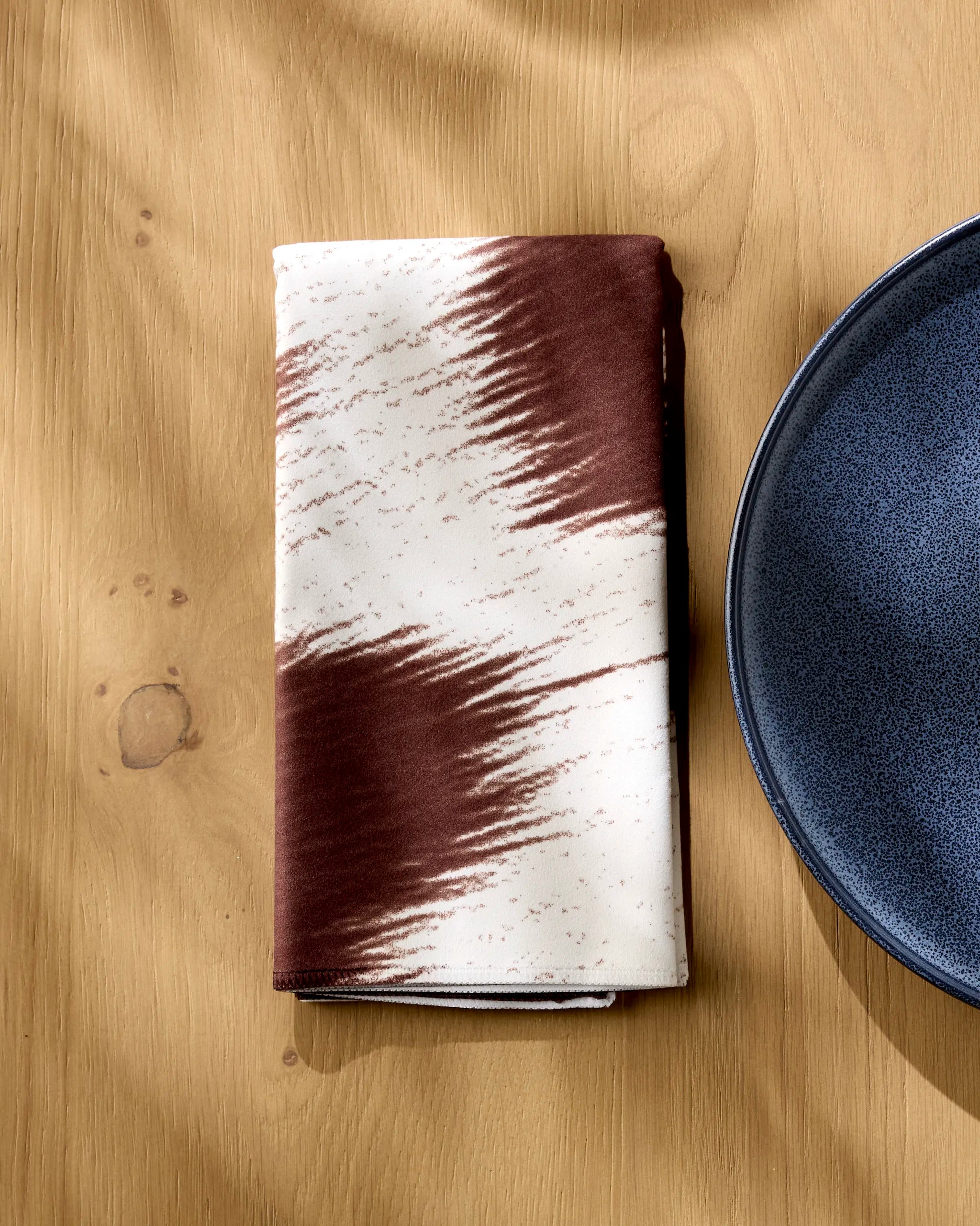 The Rodeo reusable cloth napkin by Geometry, featuring a brown and white zigzag pattern, is folded on a light wooden table beside a dark blue speckled ceramic plate. Sunlight creates gentle shadows across the setting.