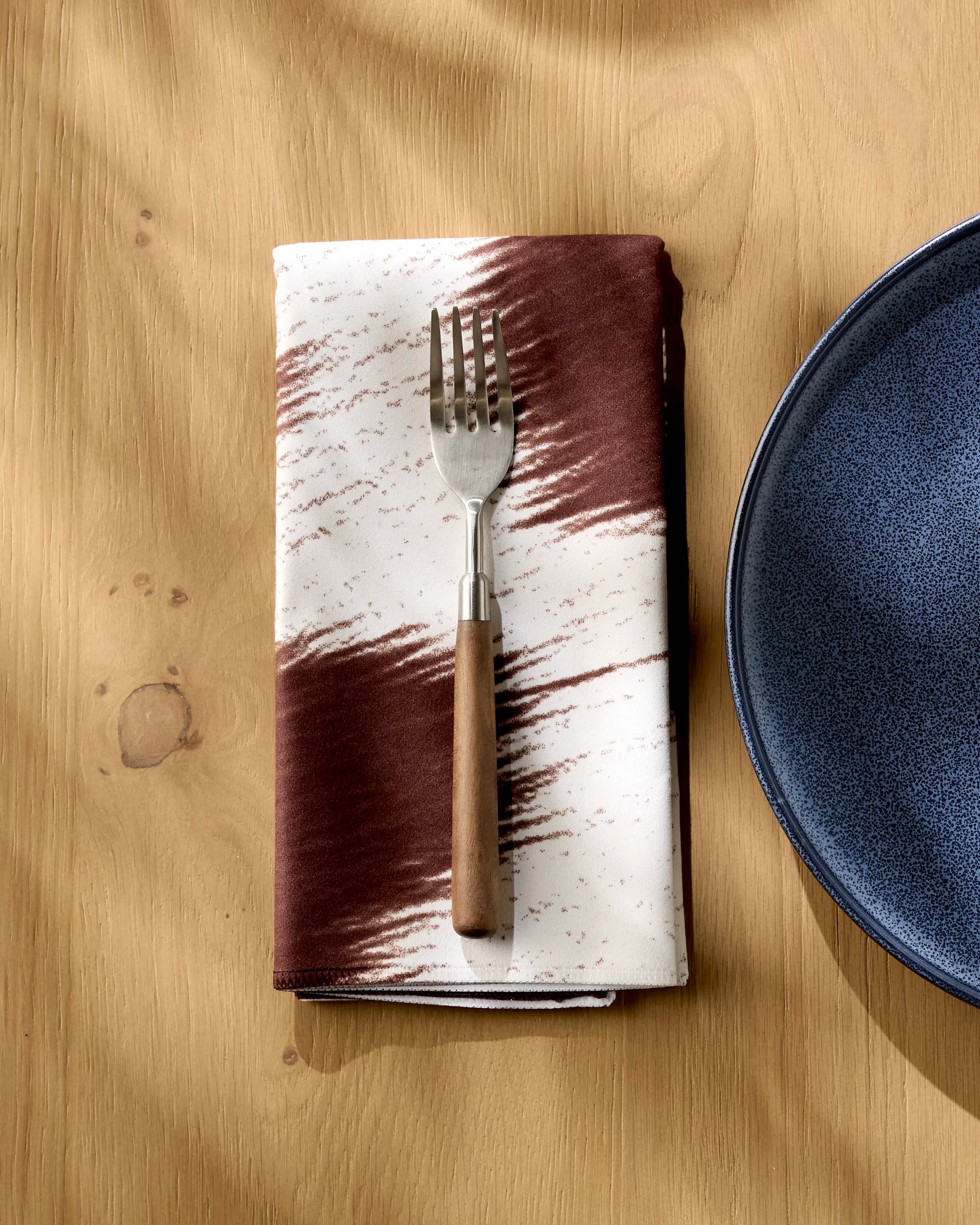 A Rodeo fork with a wooden handle rests on a brown-and-white patterned reusable Geometry napkin, beside a blue ceramic plate on a light wooden table.