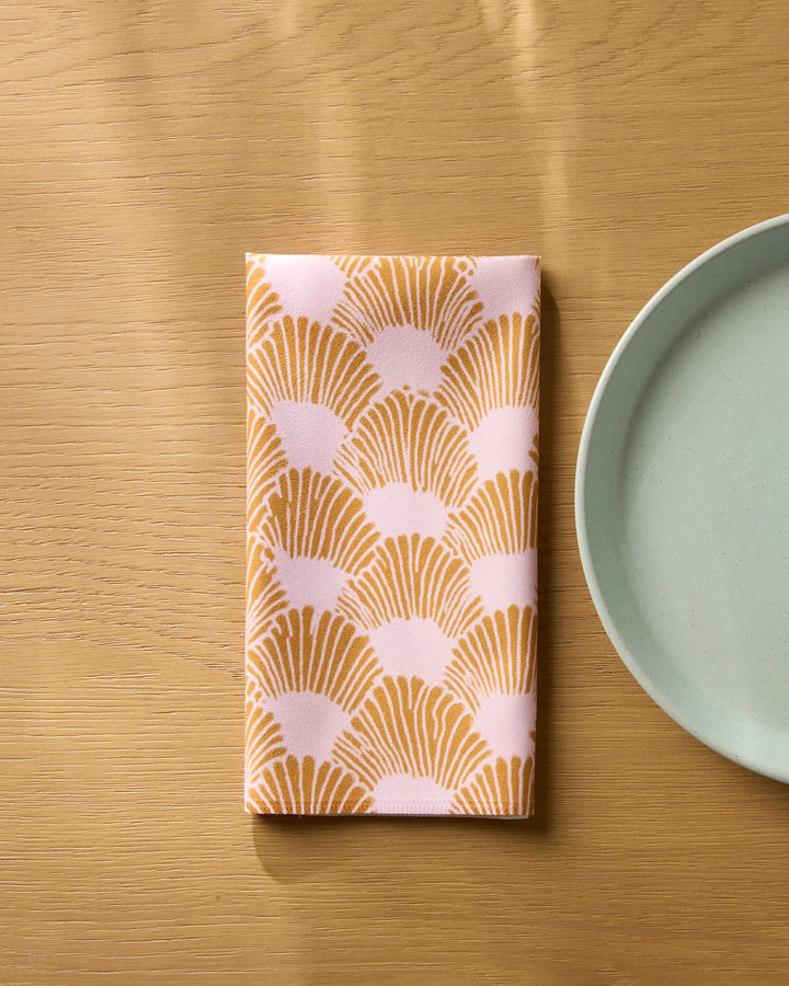 The Royal Scallop reusable napkin by Geometry, featuring an orange and pale pink fan pattern, is folded on a light wooden surface beside a light blue plate.