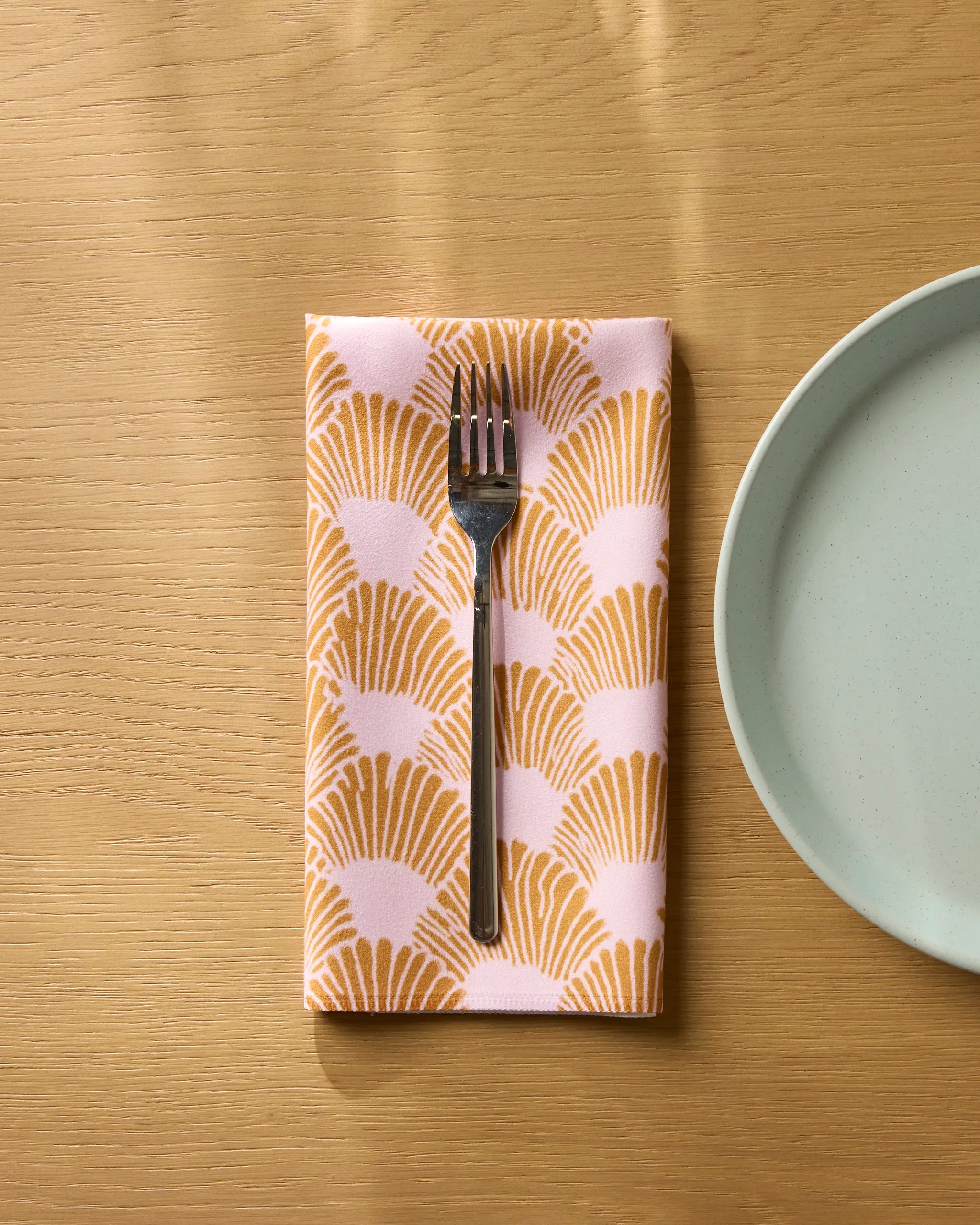 A silver fork rests on Geometry's Royal Scallop reusable napkin, featuring a pink and gold fan pattern, placed on a light wooden table beside a pale blue ceramic plate.