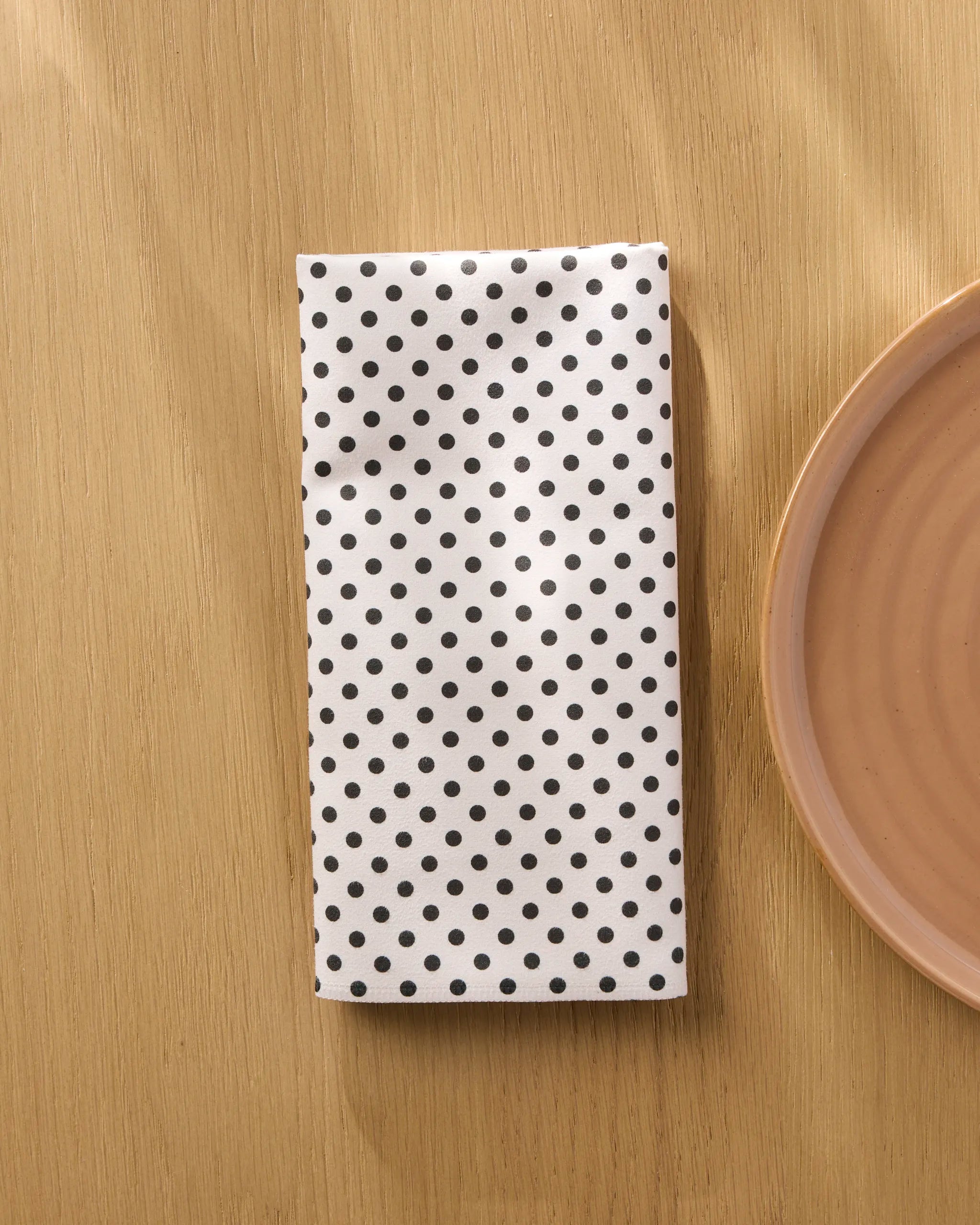The Saddle Off White recycled dinner napkin by Geometry is neatly folded on a light wood table next to the edge of a beige ceramic plate.