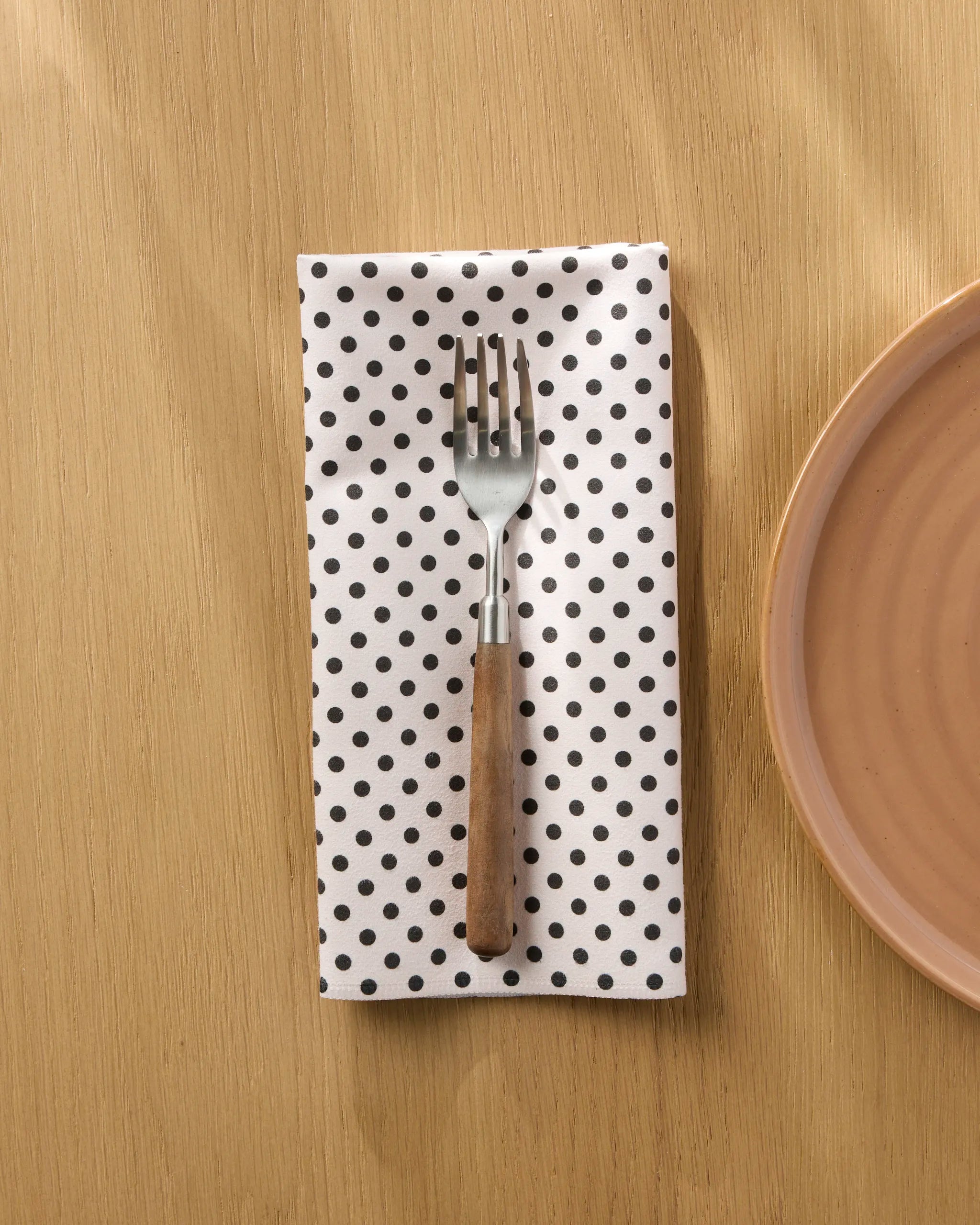 A fork with a wooden handle rests on the Geometry Saddle Off White absorbent cloth napkin with black polka dots, placed on a light wooden table beside a pink ceramic plate.