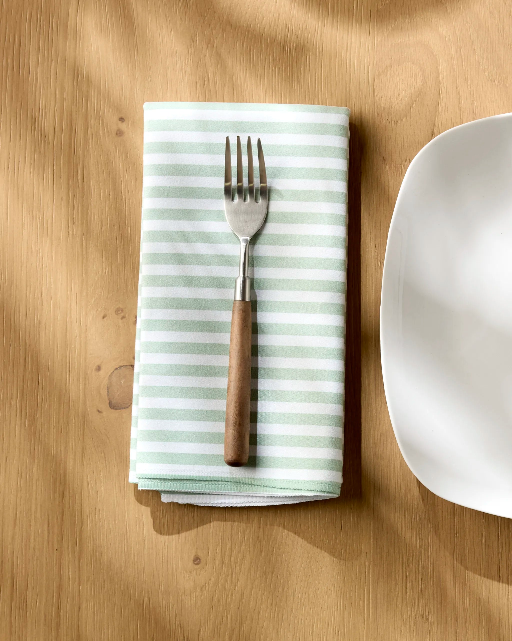 A Geometry Seagrass fork with a wooden handle rests on a folded, green-and-white striped absorbent cloth napkin beside a white, square-edged plate on a light wood table.
