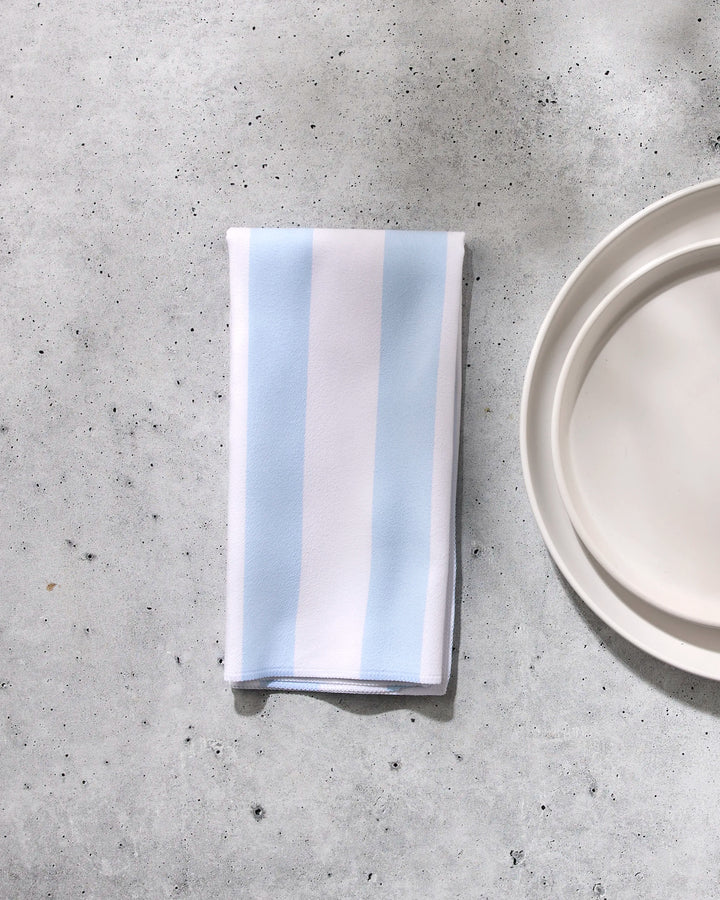 The GEOMETRY Seaside Stripes reusable dinner napkin, featuring light blue and white vertical stripes, rests on a gray textured surface beside a plain white plate, bringing style and sustainability to your table setting.