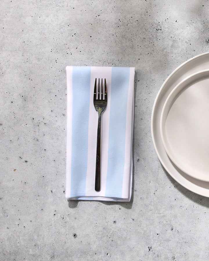 A GEOMETRY Seaside Stripes blue and white eco-friendly napkin is folded beside a silver fork and a stack of white plates on a light gray, textured surface.