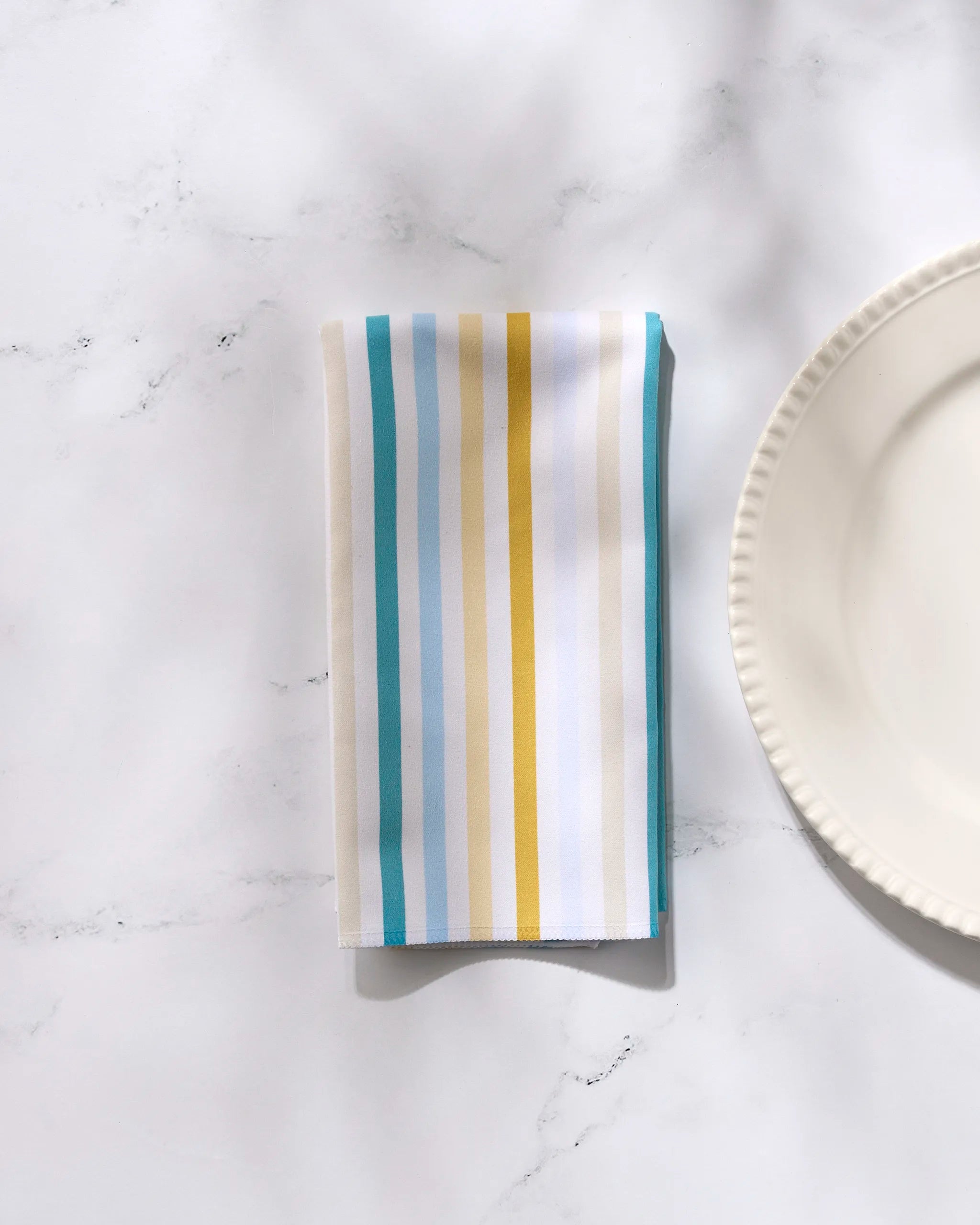 The GEOMETRY Seaside Sunshine Dinner Napkin Set features a folded reusable napkin with blue, yellow, and white vertical stripes beside a scalloped white plate on marble, perfect for stylish table settings.