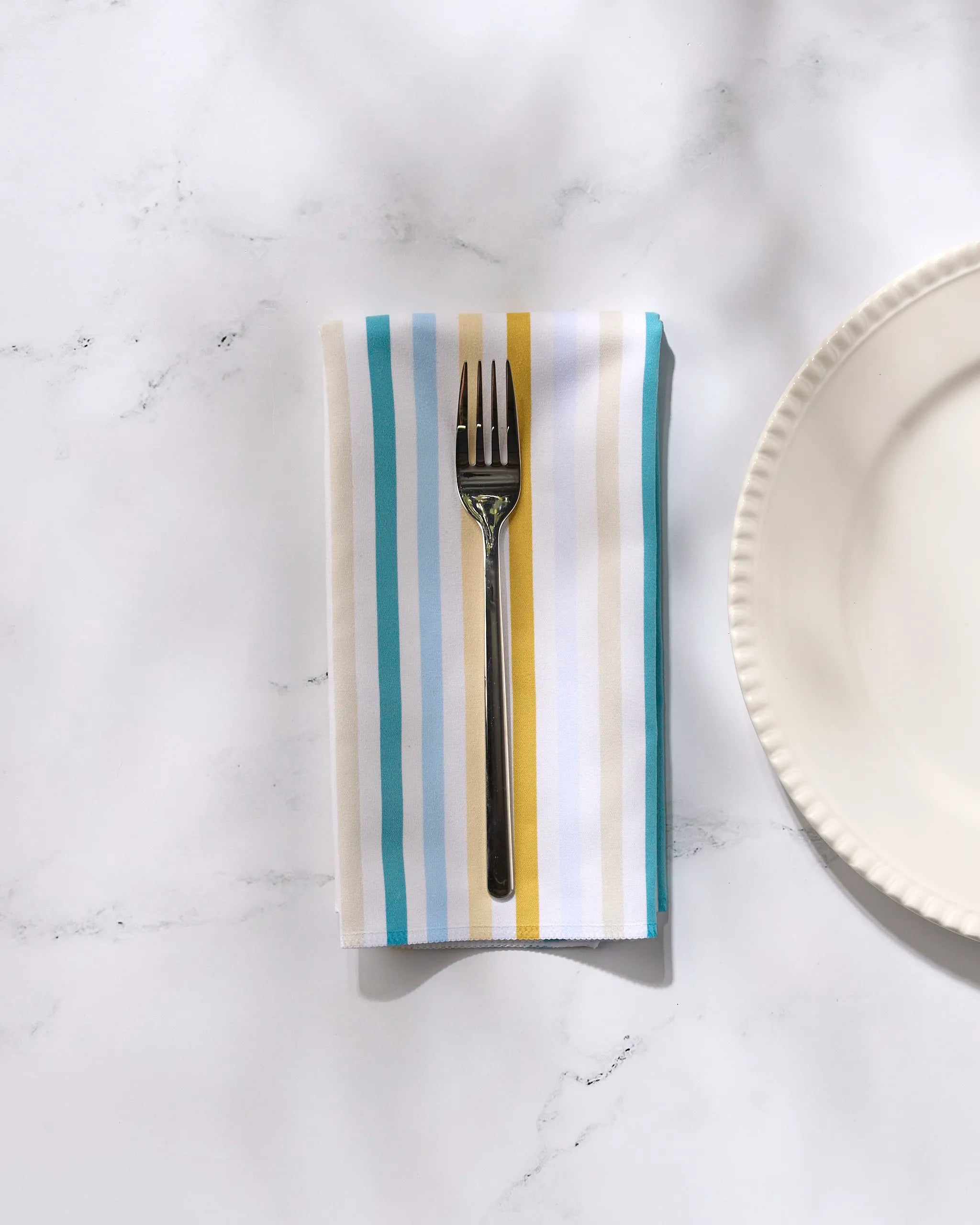 A metal fork sits on the GEOMETRY Seaside Sunshine reusable napkin, featuring blue, yellow, and white stripes, next to a white scalloped plate on a marble surface.