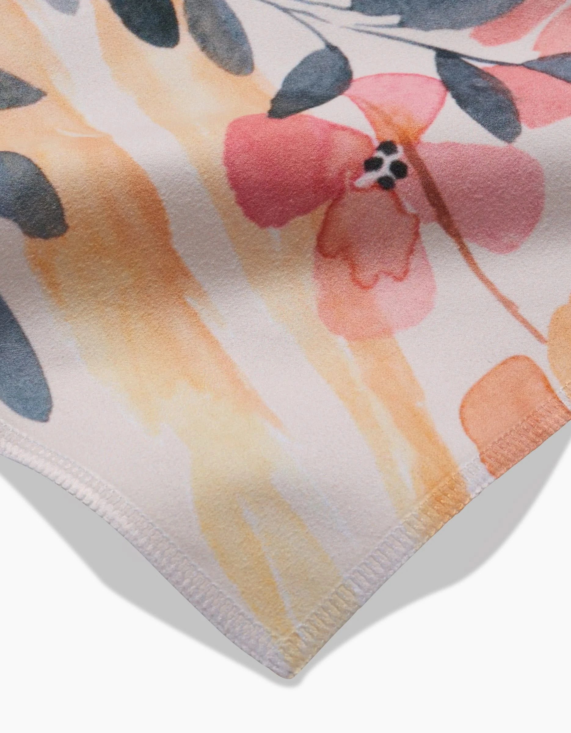 A close-up of GEOMETRY’s Sleepy Fields Geosuede shows a watercolor floral pattern in pink, peach, and orange with green leaves. The fabric edge is neatly stitched on a light background.