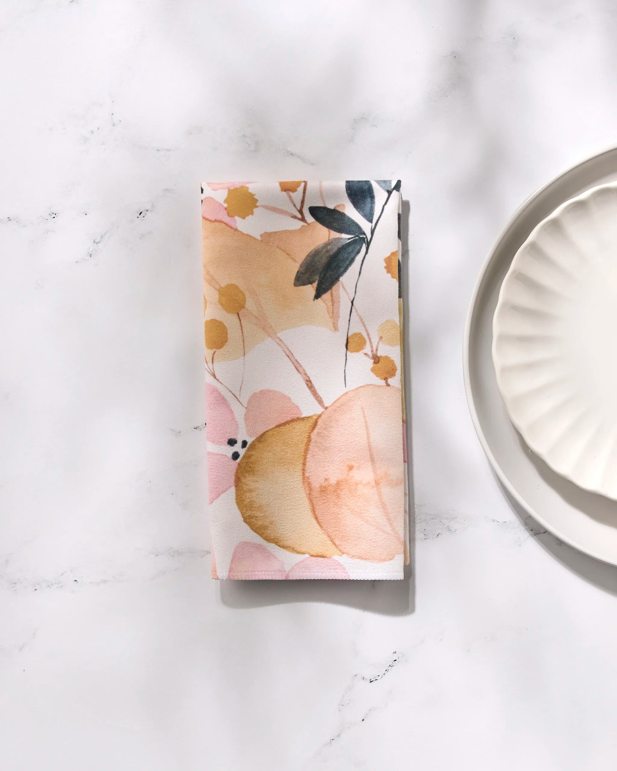 The GEOMETRY "Sleepy Fields" reusable napkin, featuring a soft floral print in pastel pink, peach, and yellow, rests on marble by a white scalloped plate—a perfect touch for your dinner napkin set.