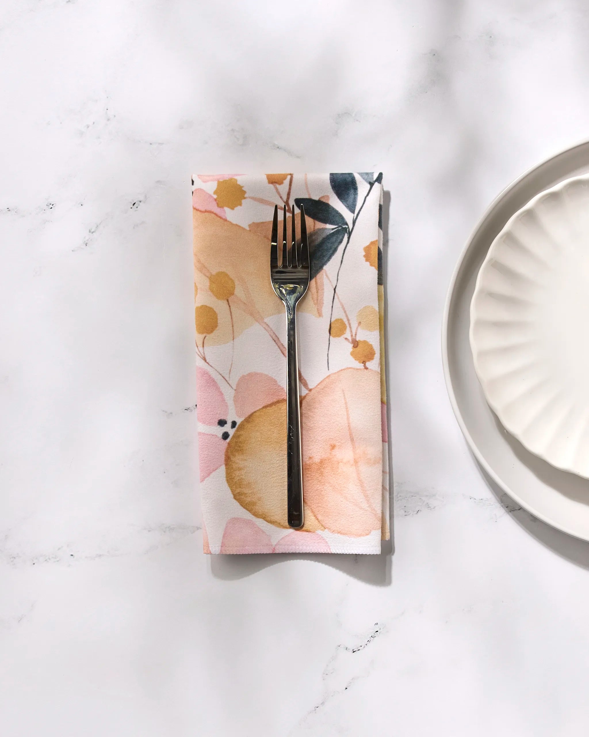 A fork rests on the GEOMETRY Sleepy Fields floral-patterned recycled fabric napkin atop a white marble surface beside a scalloped white plate, creating an elegant, minimal table setting in soft shadows and bright light.