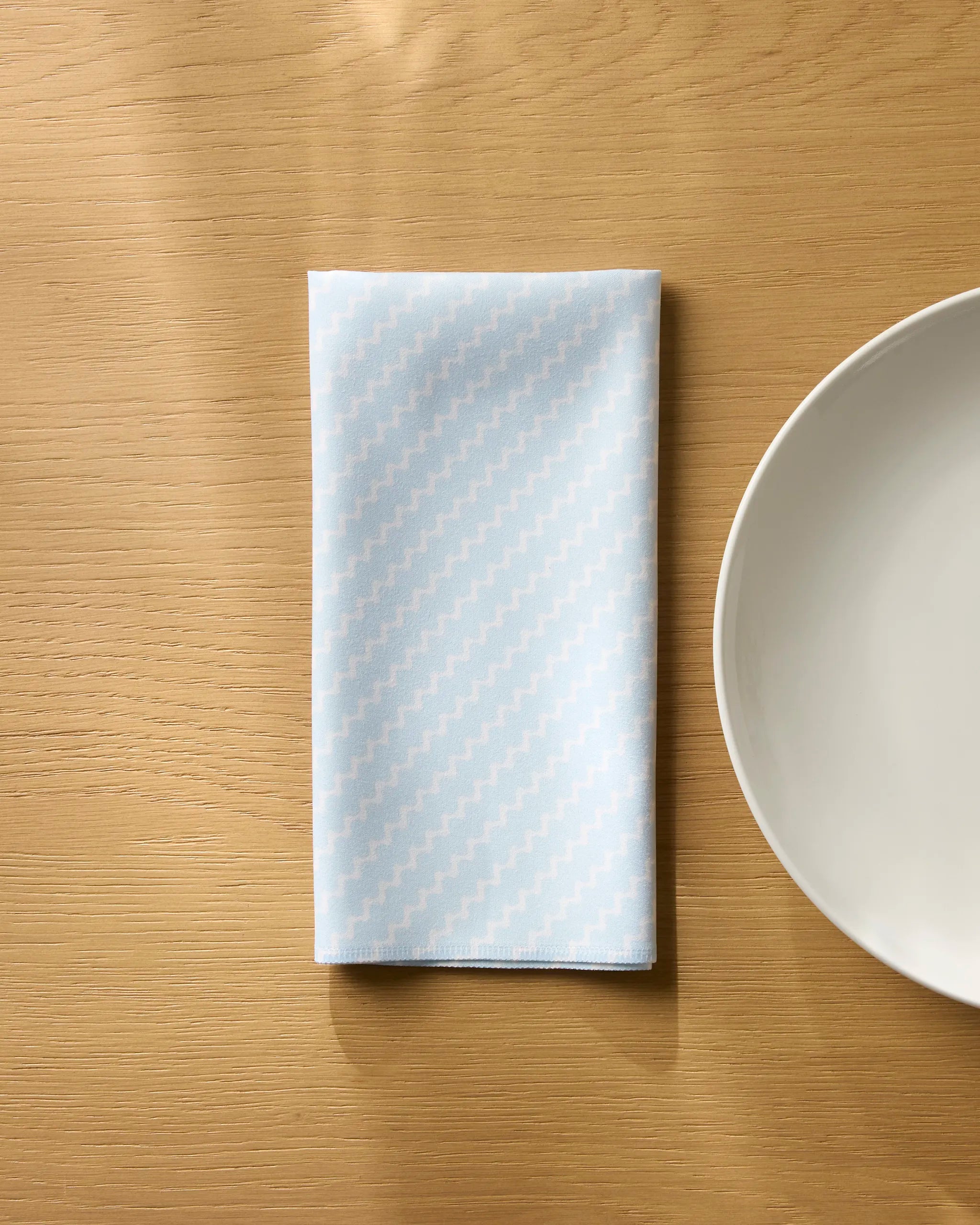 A Soft Fringe reusable napkin by Geometry, in light blue with a subtle zigzag pattern, is neatly folded on a wooden table beside an empty white plate.