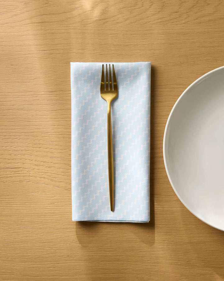 A gold fork rests on Geometry’s Soft Fringe light blue reusable napkin with a zigzag pattern, folded on a wooden table next to an empty white plate.
