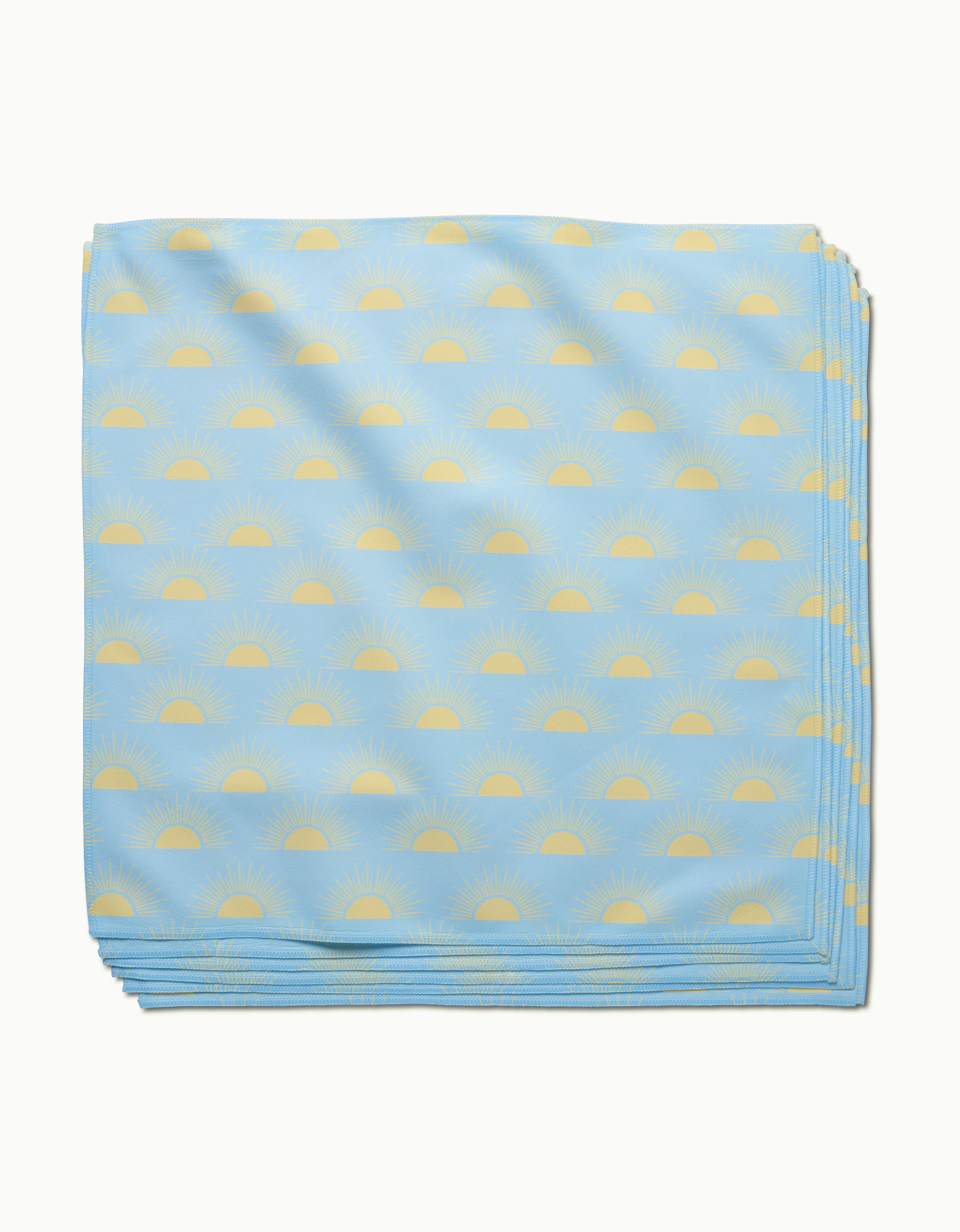 The GEOMETRY Soft Summer Sun dinner napkin, folded from light blue Geosuede fabric with a minimalist pattern of yellow sunrises and radiating rays, is neatly stacked against a white background.