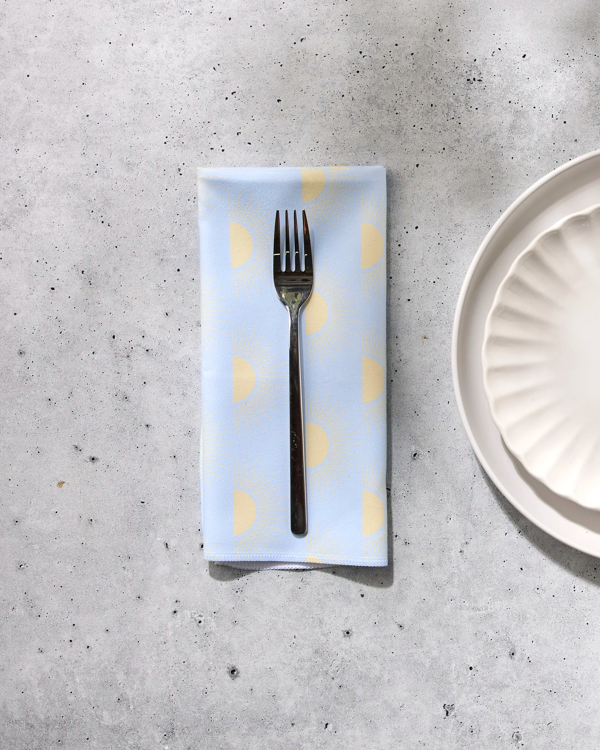 A fork rests on GEOMETRY’s "Soft Summer Sun" blue reusable cloth napkin with yellow sun patterns, placed on a gray speckled surface next to a white, scalloped-edge plate.