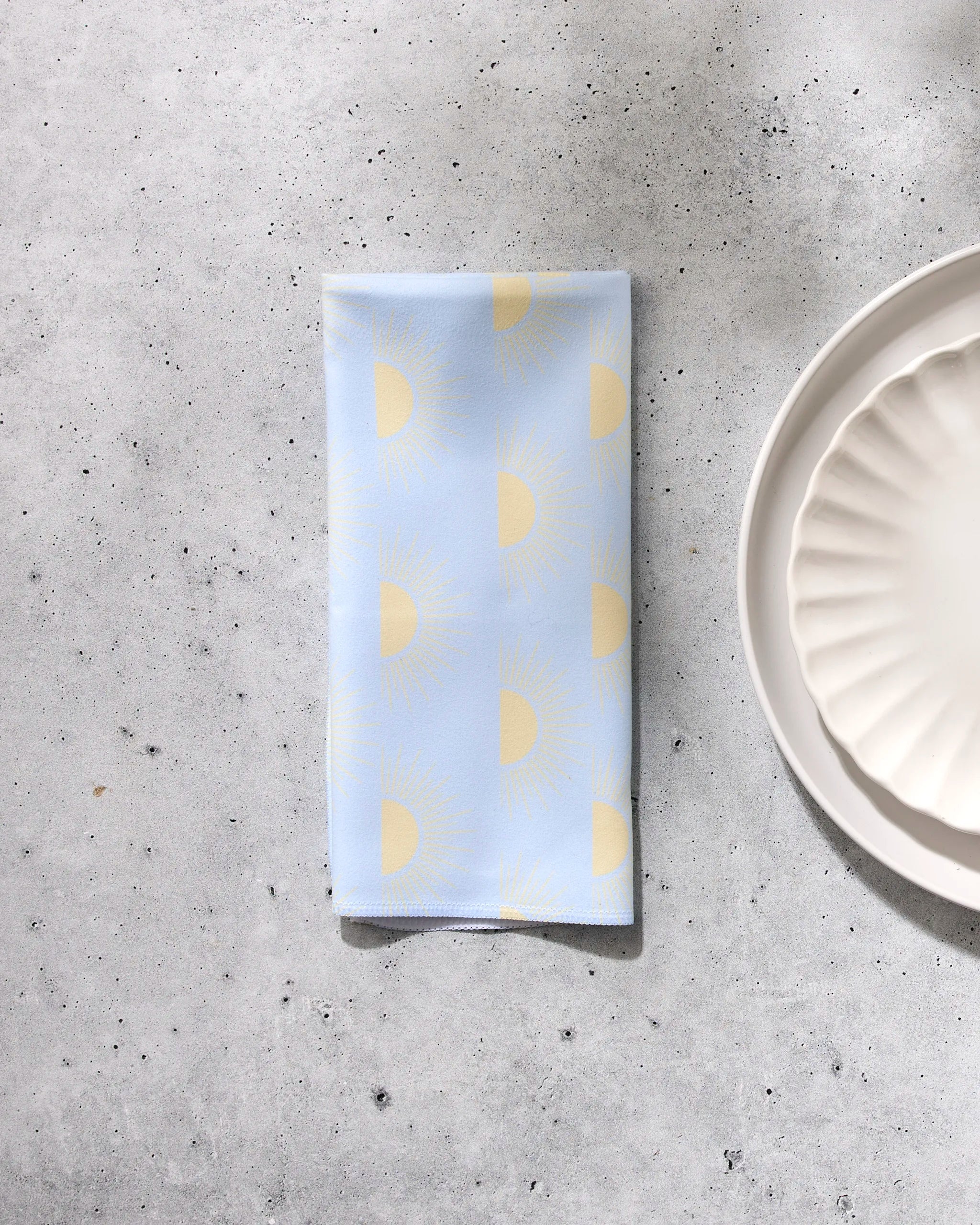 The GEOMETRY "Soft Summer Sun" reusable cloth napkin, featuring a yellow sun pattern on light blue fabric, is folded on a speckled gray surface beside a white scalloped ceramic plate.