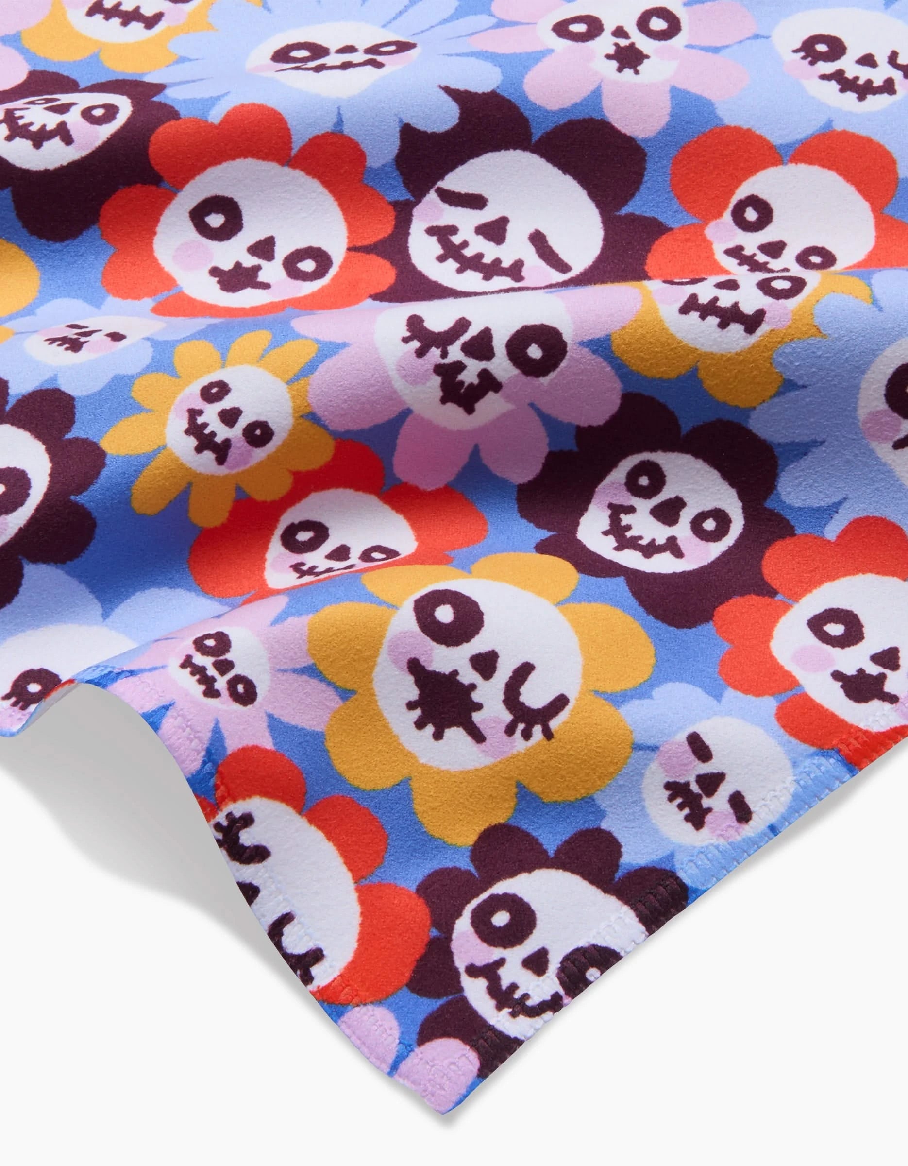 The So Lazo Spooky Bouquet Geosuede fabric showcases cartoon skulls with flower petals in red, yellow, pink, and purple on light blue—its vibrant, playful design makes it perfect for quirky dinner napkins.