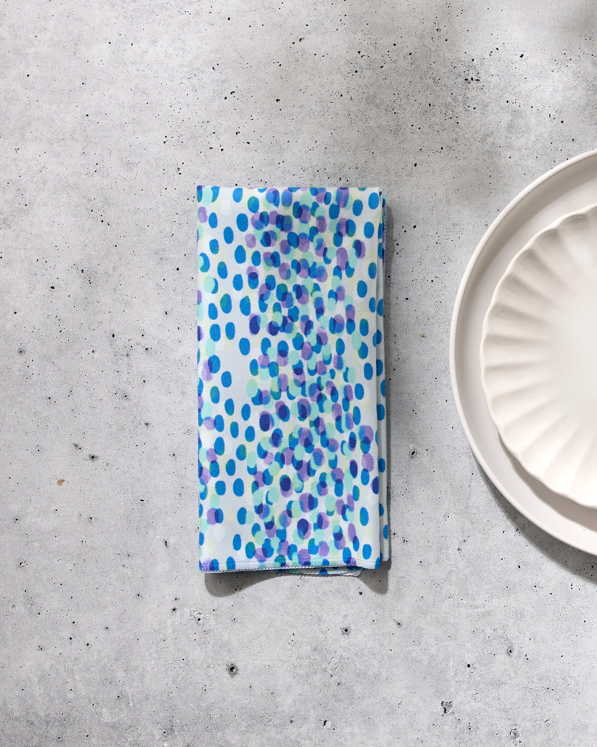 The Spotty Summer napkin by GEOMETRY, featuring blue and purple polka dots on white recycled fabric, rests on a light gray textured surface beside a scalloped-edge plate, bringing sustainable charm to your table setting.