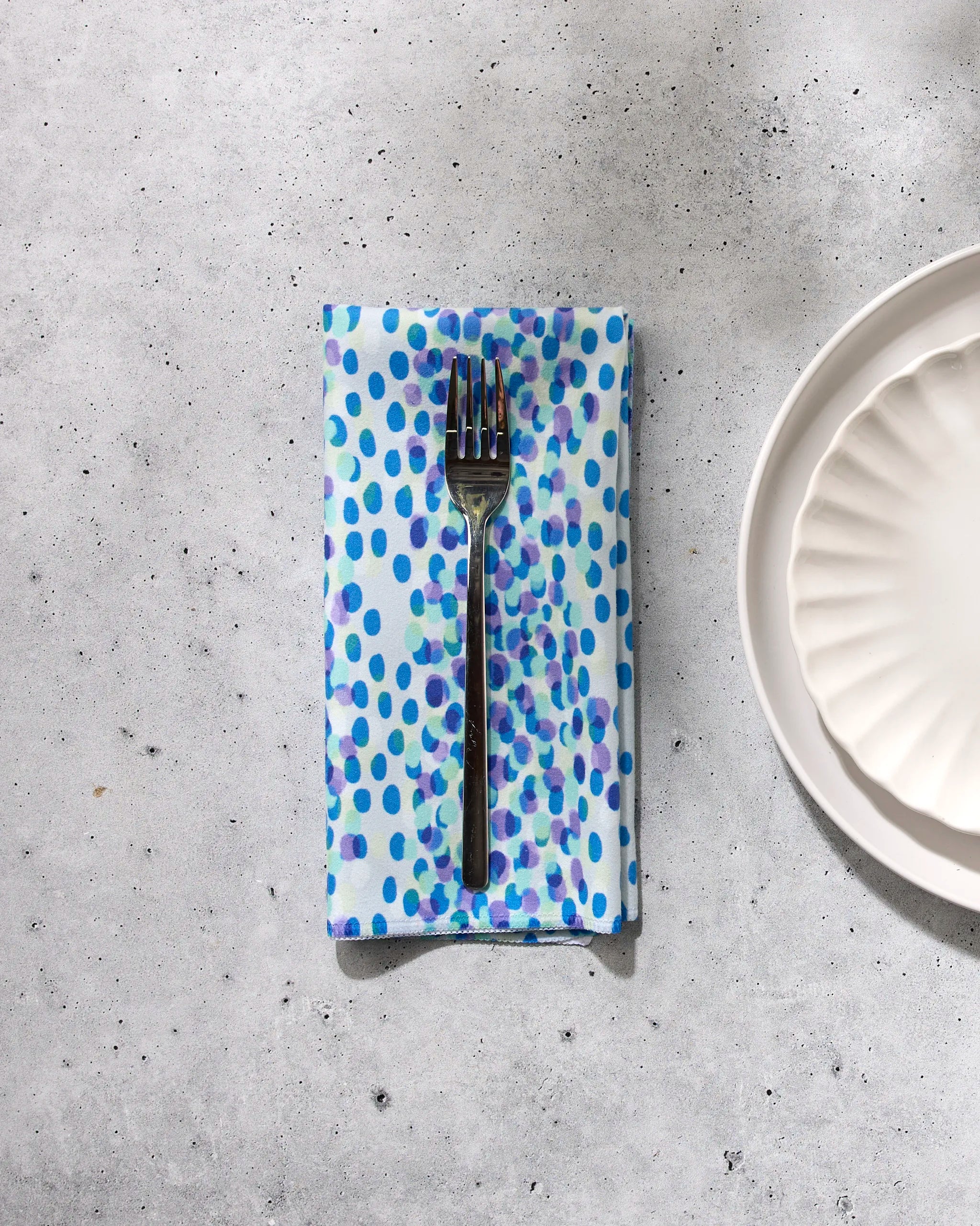 A fork rests on the GEOMETRY Spotty Summer blue and purple polka dot dinner napkin set, made from sustainable table linens, placed on a light gray surface beside a white scalloped plate.