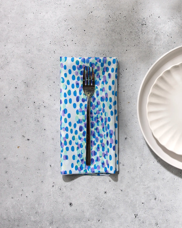 A fork rests on the GEOMETRY Spotty Summer blue and purple polka dot dinner napkin set, made from sustainable table linens, placed on a light gray surface beside a white scalloped plate.