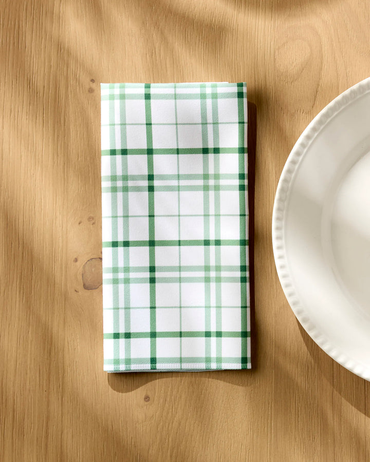 The Hufton Studio Spring Picnic Plaid, a folded white reusable table napkin with green and dark green plaid, rests on a light wooden table beside a white scalloped plate as soft sunlight casts gentle shadows.