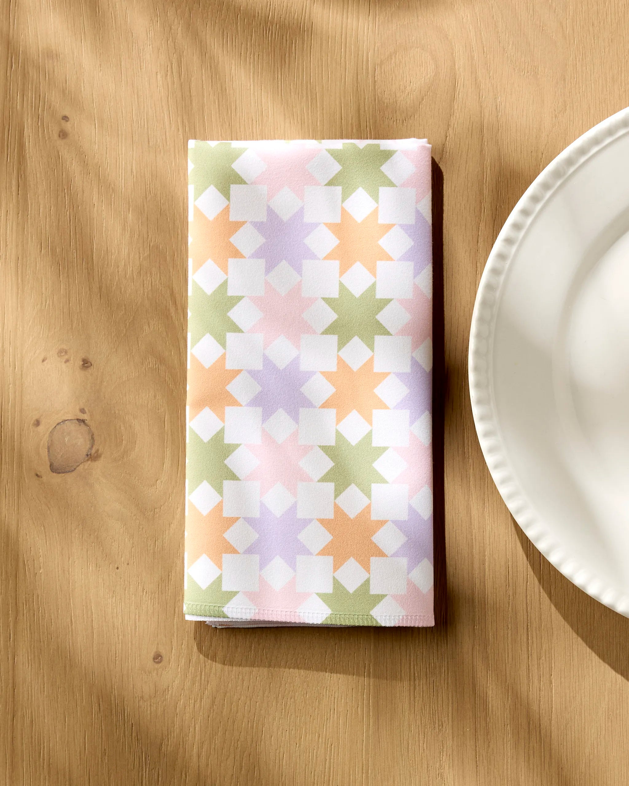 A folded Kayla Ann Spring Quilt napkin with a pastel geometric pattern in green, pink, purple, and orange rests beside a white plate on a wooden table.
