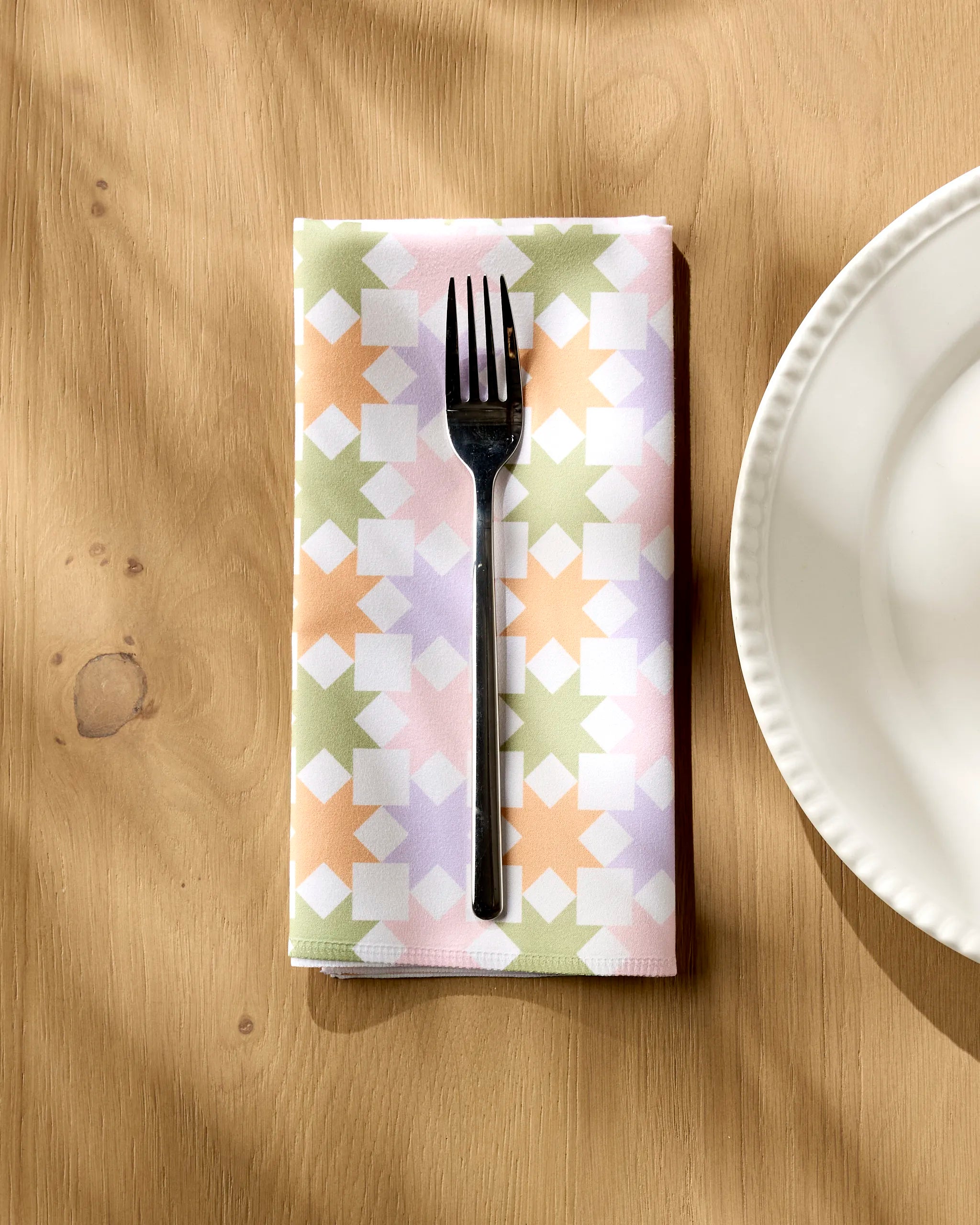 A silver fork rests on the Kayla Ann Spring Quilt, a pastel-colored, soft absorbent napkin with a geometric pattern, placed on a wooden table beside the edge of a white plate.