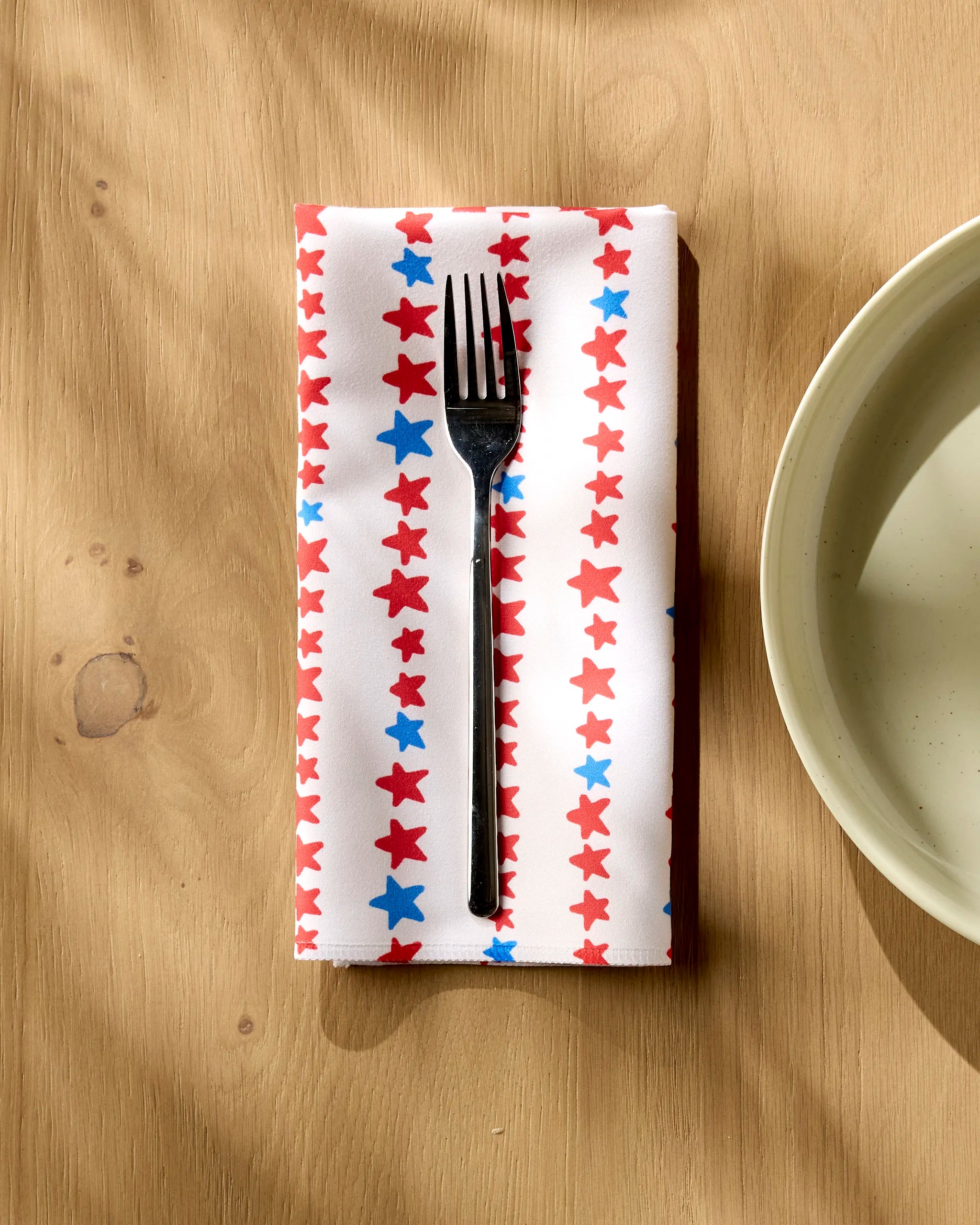 A fork rests on Kayla Ann’s Starry Stripes eco-friendly napkin, adorned with red and blue stars, on a wooden table beside a pale plate. Sunlight casts gentle shadows over the setting.