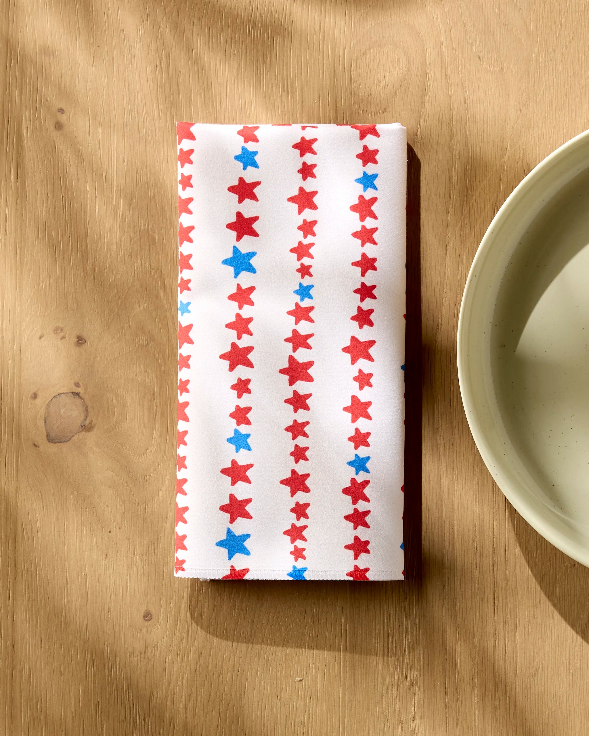A folded Kayla Ann Starry Stripes eco-friendly napkin, featuring vertical red and blue stars, rests on a wooden table beside a beige plate.