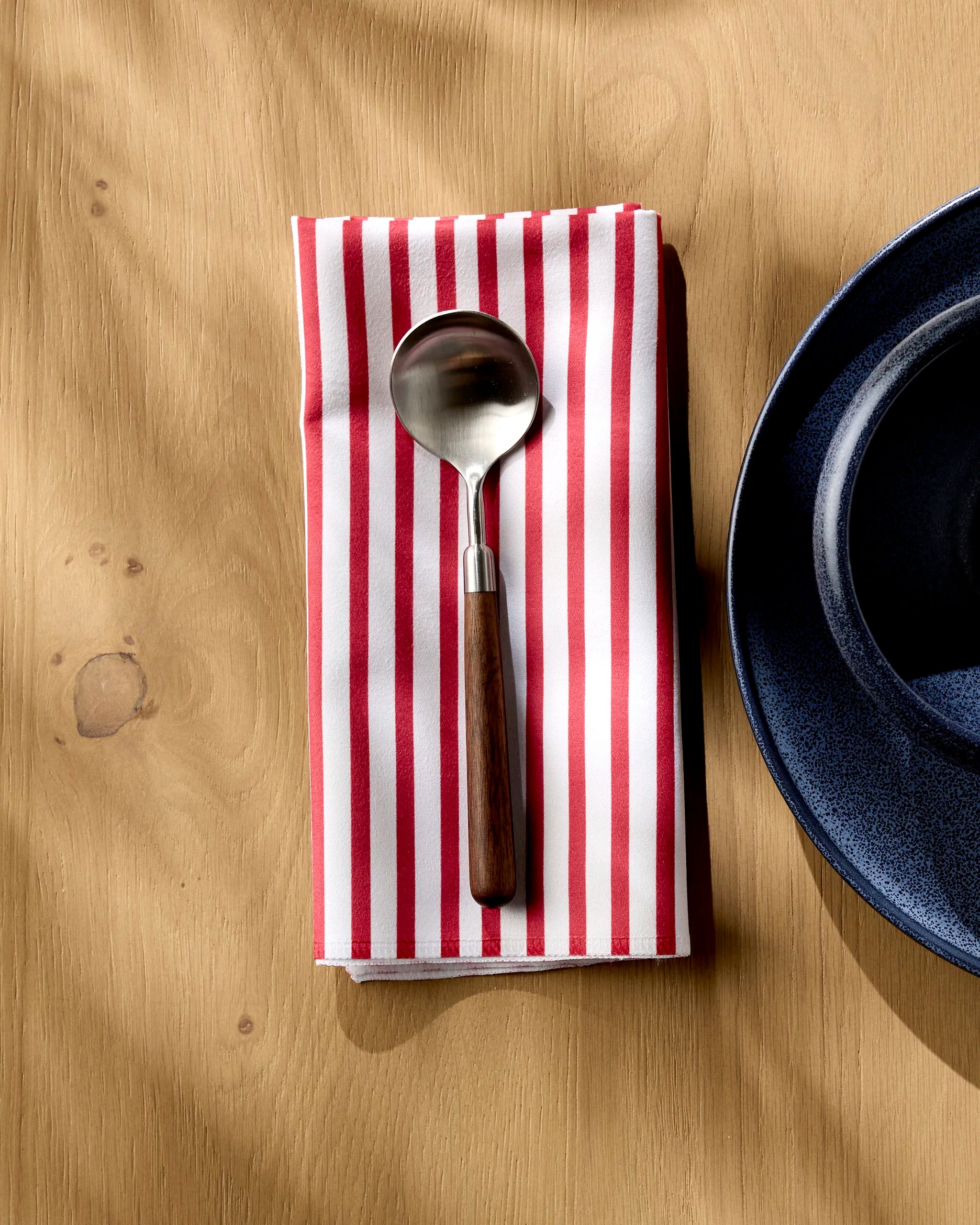 The Mindfulnice Stars And Stripes metal spoon with a wooden handle rests on a red and white striped reusable cloth napkin beside a dark blue ceramic plate on a light wooden table.
