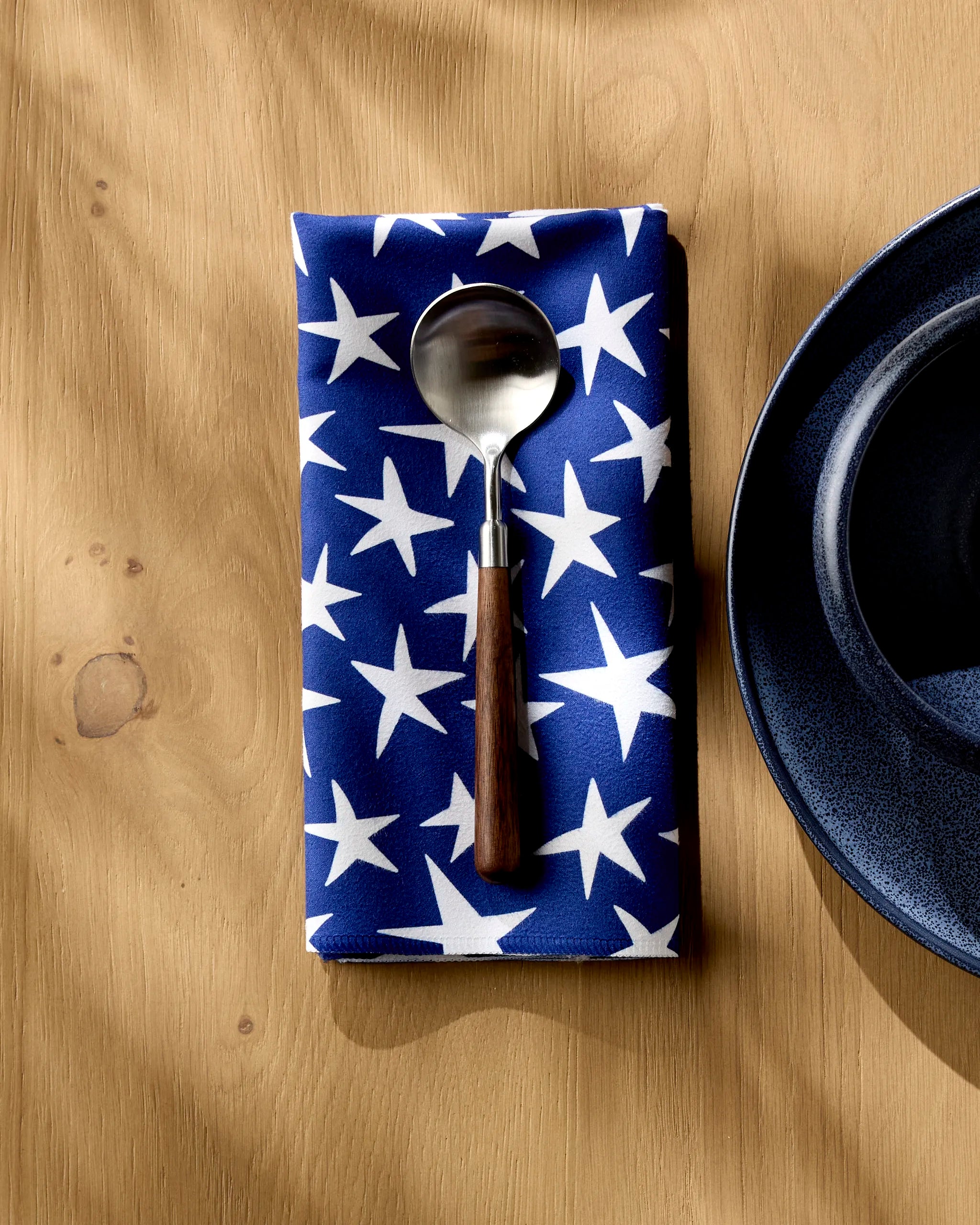 A spoon rests on the Mindfulnice Stars And Stripes blue reusable cloth napkin with white stars, set on a wooden table beside a dark blue plate.