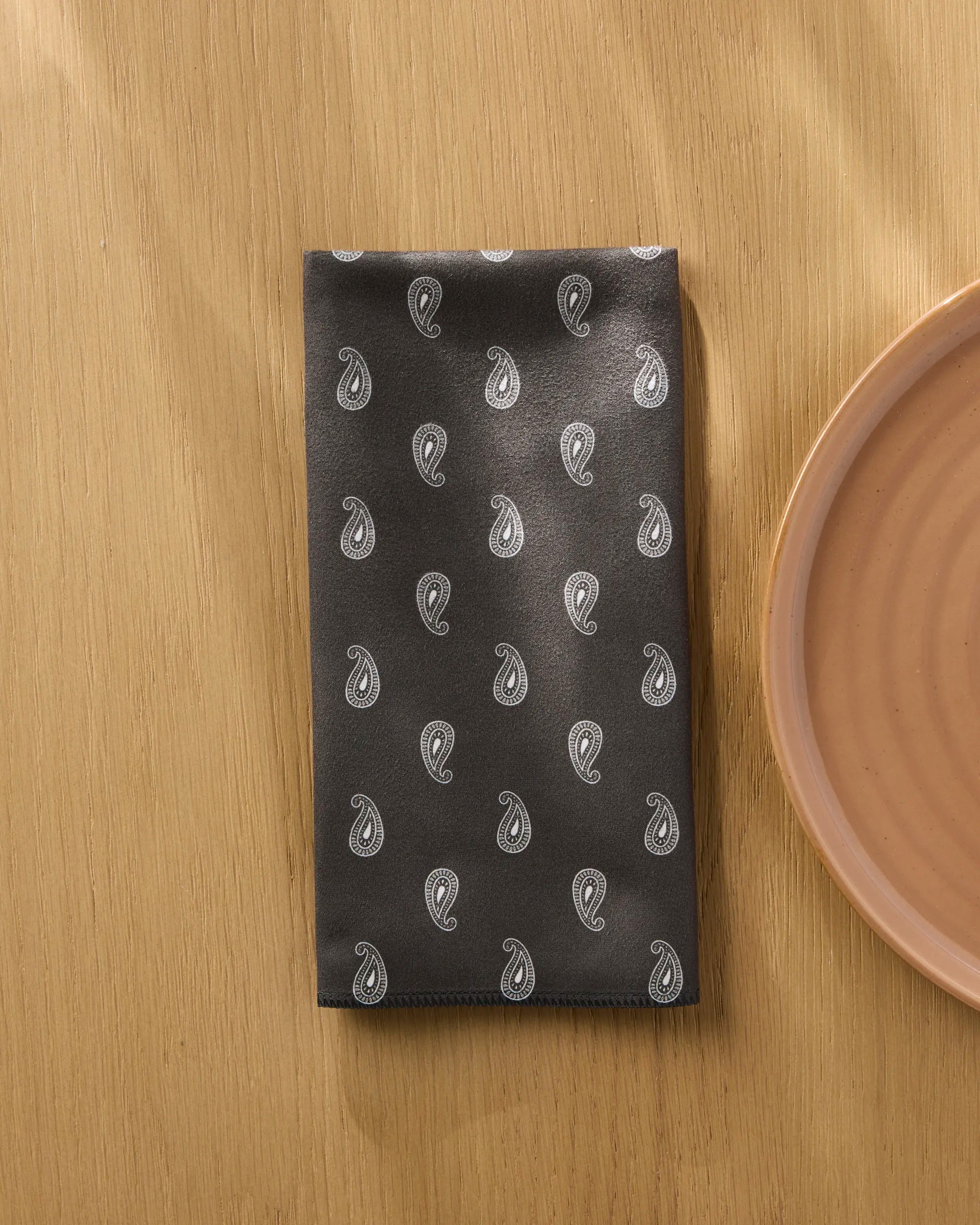 A black Stirrup Espresso napkin by Geometry with a white paisley pattern rests on wood beside a round peach ceramic plate.