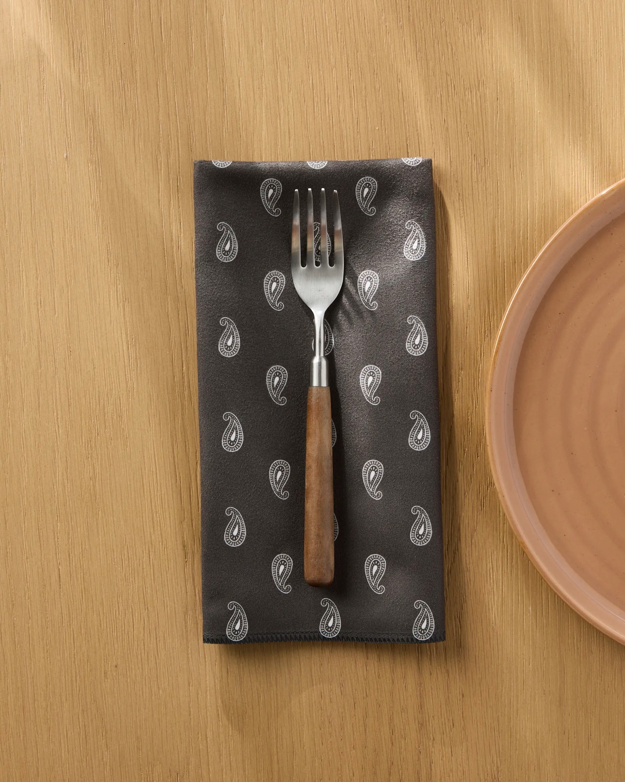 The Geometry Stirrup Espresso napkin with a dark paisley pattern rests on a light wood table, paired with a fork and next to a peach-colored ceramic plate.