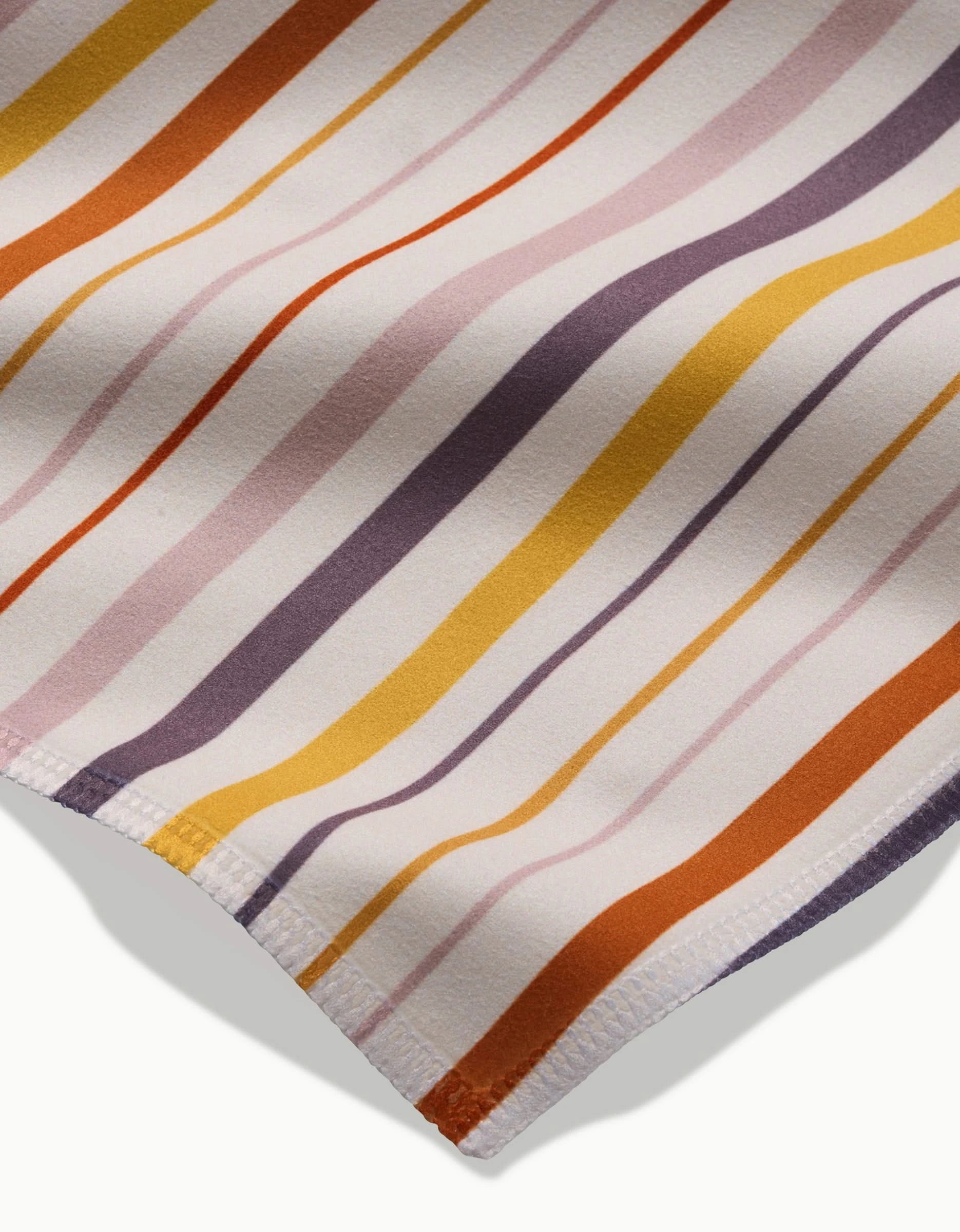 Close-up of GEOMETRYs Stripe Season dinner napkins: Geosuede fabric with diagonal orange, gold, lavender, and purple stripes on white. Made from recycled materials, features smooth texture and neatly stitched edges.