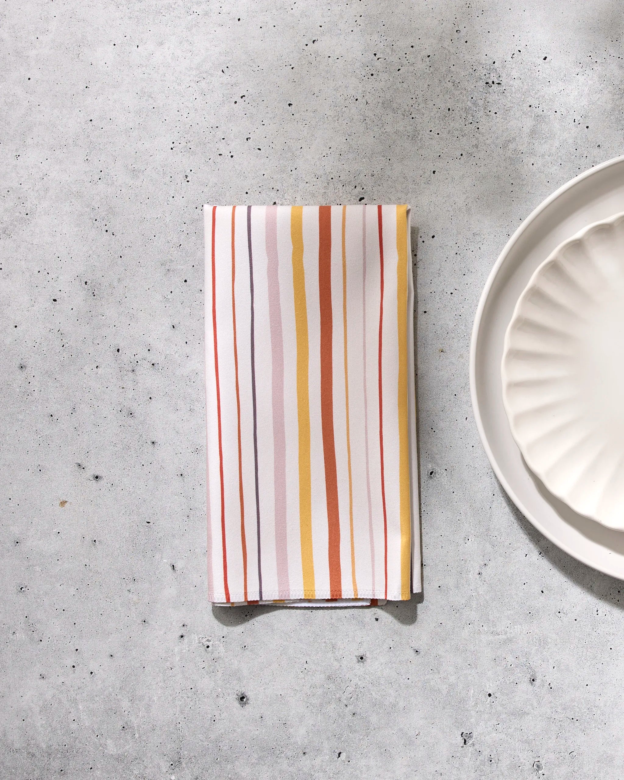 The GEOMETRY Stripe Season folded reusable napkin featuring vertical orange, yellow, and brown stripes rests on a light gray speckled surface beside the edge of a white scalloped plate.