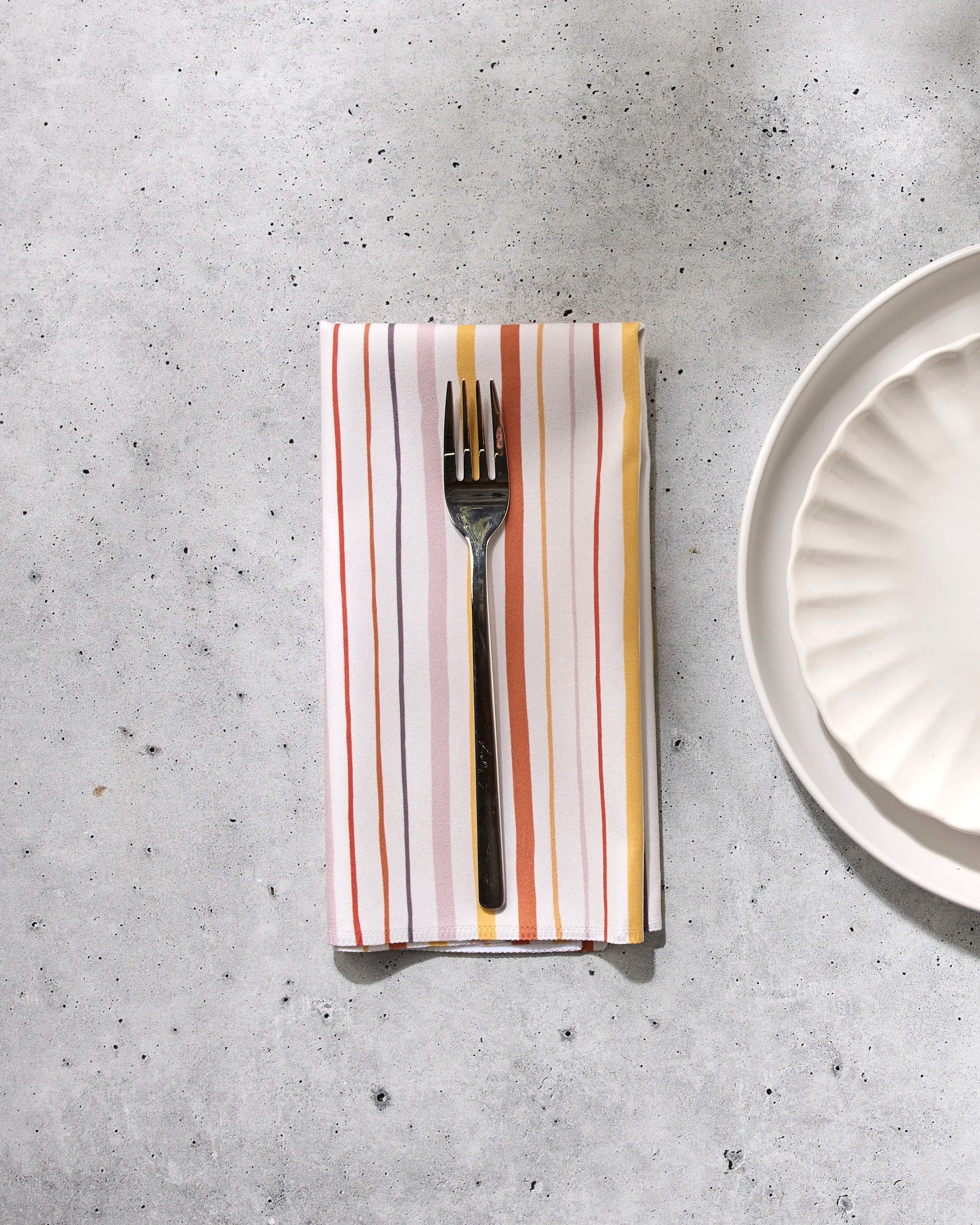 A fork rests on the GEOMETRY Stripe Season reusable dinner napkin with orange, red, and yellow stripes, placed beside a white scalloped-edge plate on a light gray speckled surface.