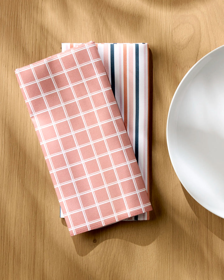 Two neatly folded Striped Harvest napkins by Charlie Rowan Designs are placed side by side on a wooden table next to a white ceramic plate.