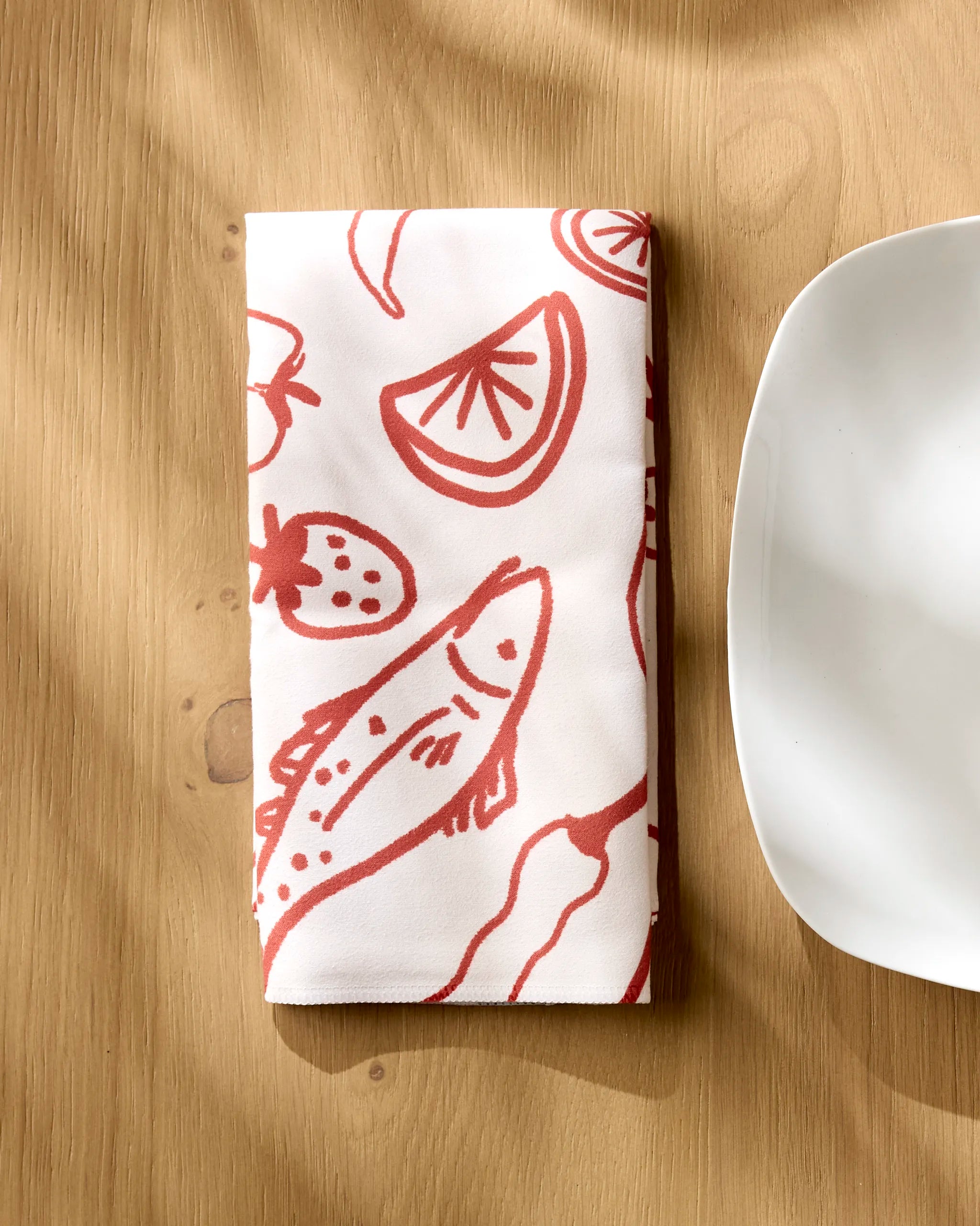 A folded white "Summer Air" cloth napkin by Geometry, adorned with red illustrations of a fish, lemon wedge, strawberry, and vegetables, rests on a wooden table beside a white plate.