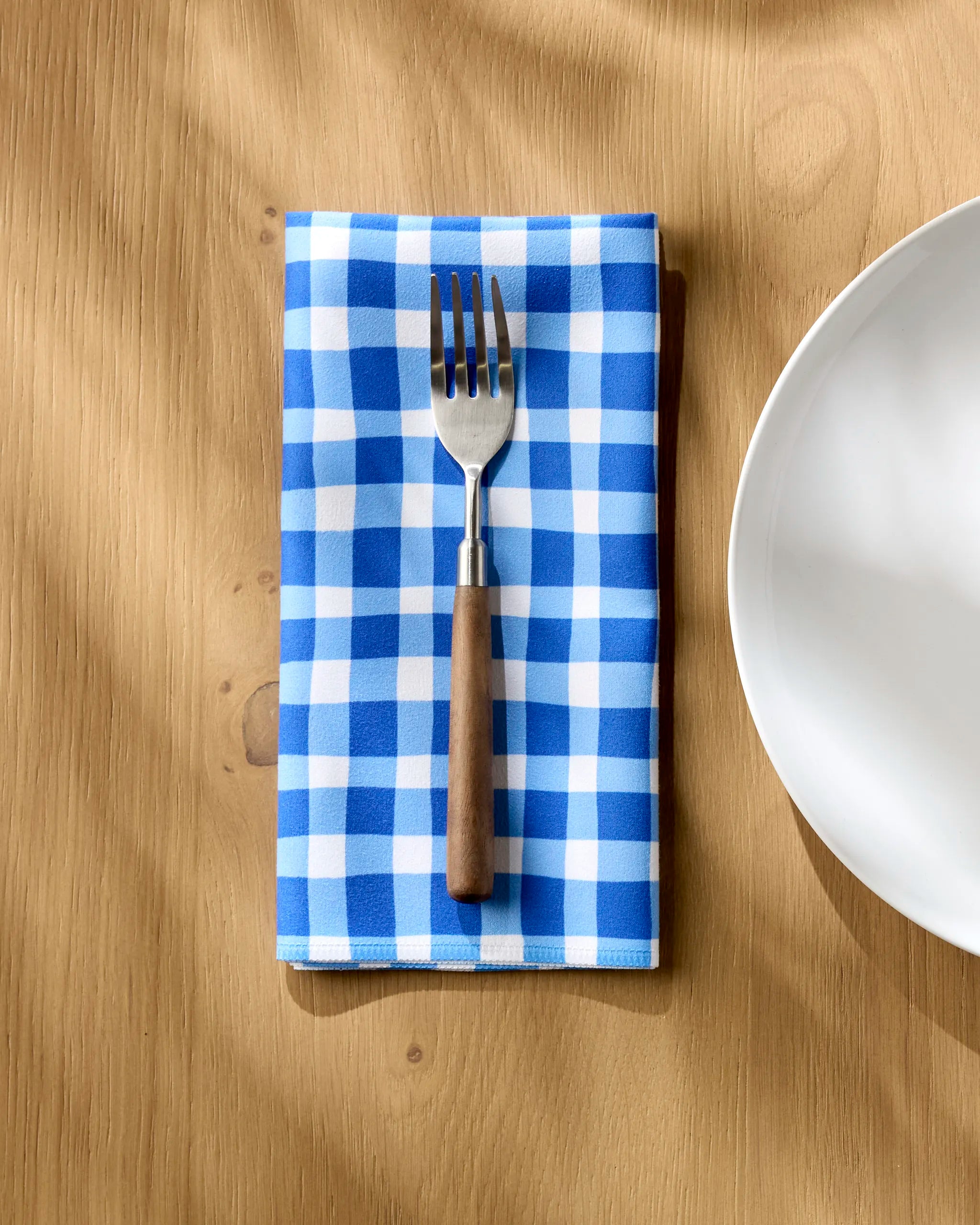 A fork with a wooden handle rests on a folded Danelys Sidron Summer Plaid blue and white checkered napkin beside a white plate on a light wood table.
