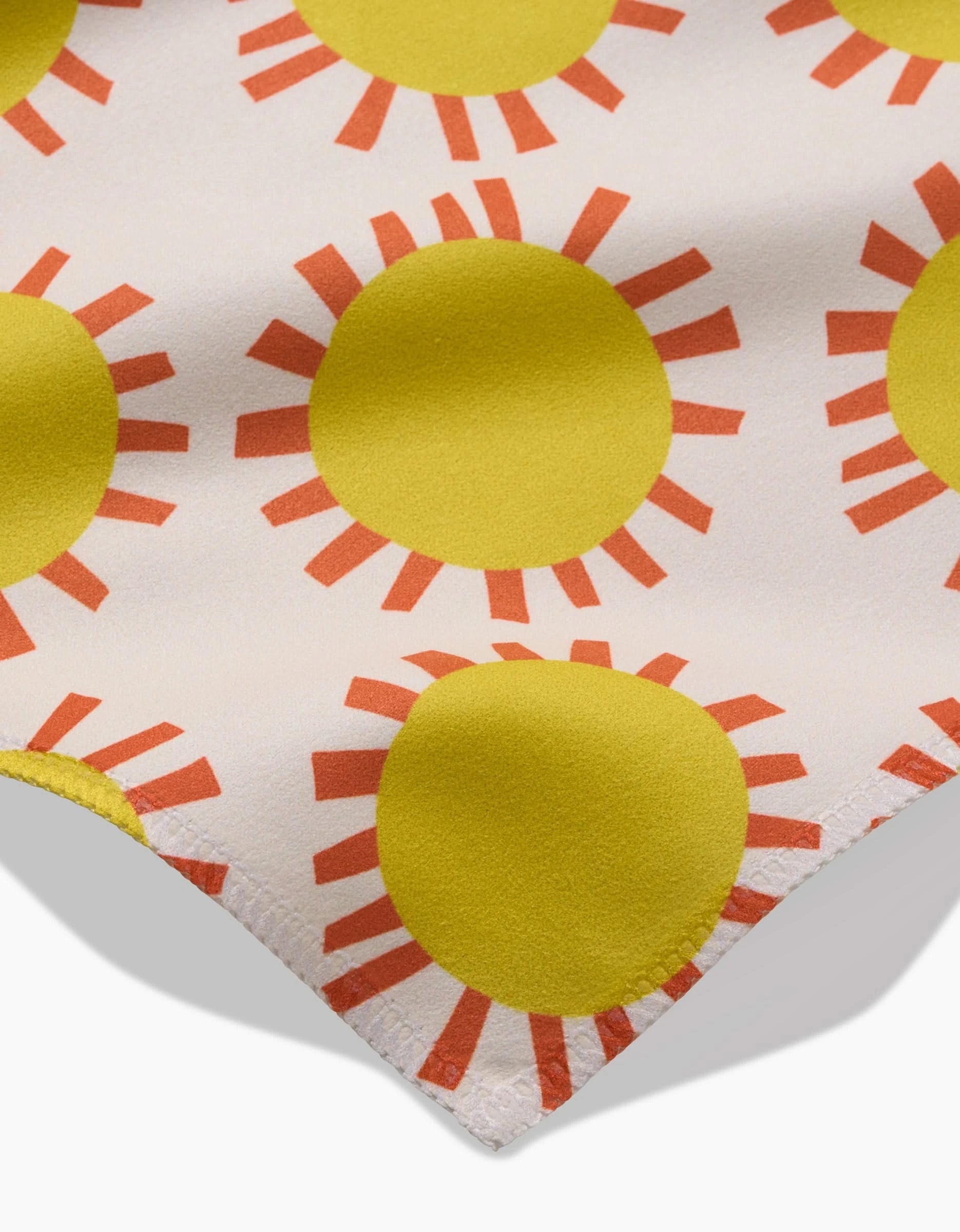A close-up of Lizzie Clarks Sunny Day Geosuede fabric shows yellow suns with red-orange rays on white. A stitched edge suggests it could be an eco-friendly Dinner Napkin made from post-consumer recycled materials.