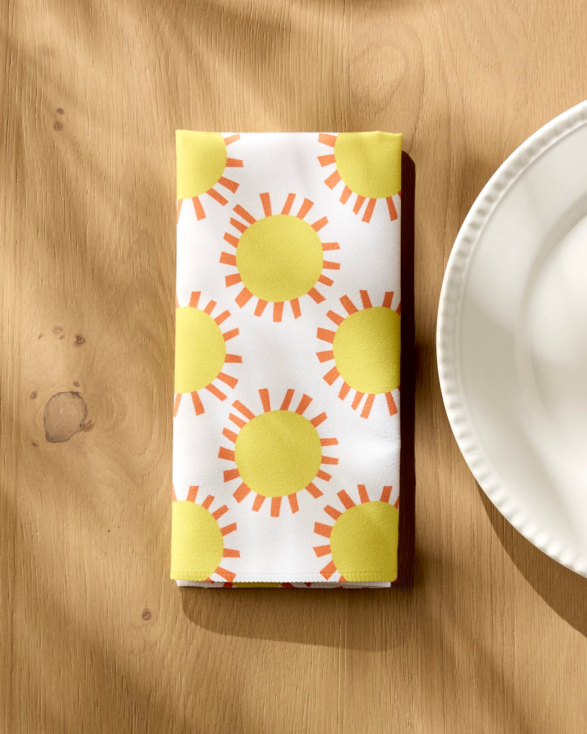 The Lizzie Clark "Sunny Day" white reusable napkin, featuring yellow suns and orange rays, lies folded on a light wooden table beside a white plate, with sunlight and gentle shadows illuminating the scene.