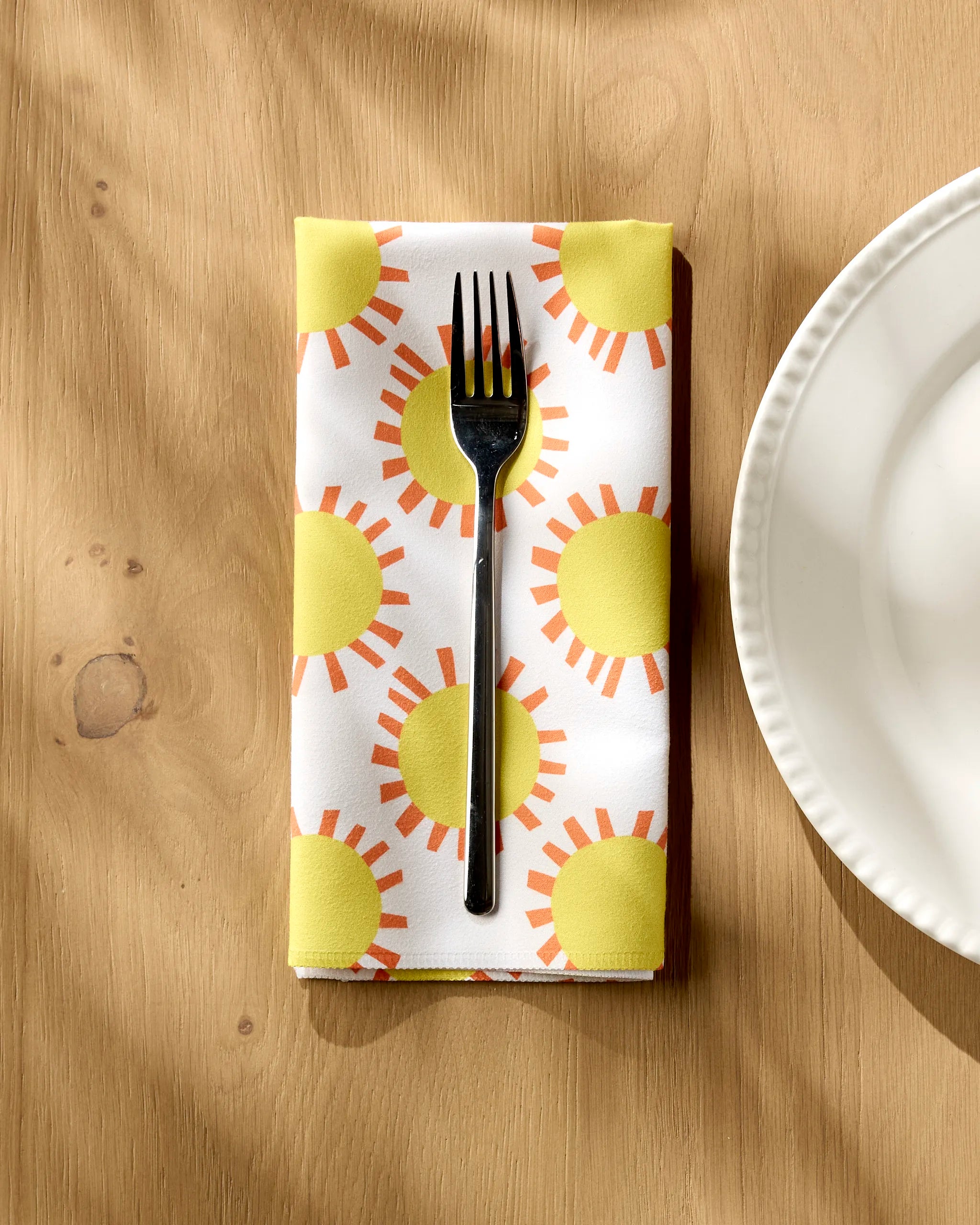 A silver fork sits on Lizzie Clark’s Sunny Day reusable cloth napkin, featuring a bright yellow and orange sun pattern, on a wooden table beside a white, round-edged plate.