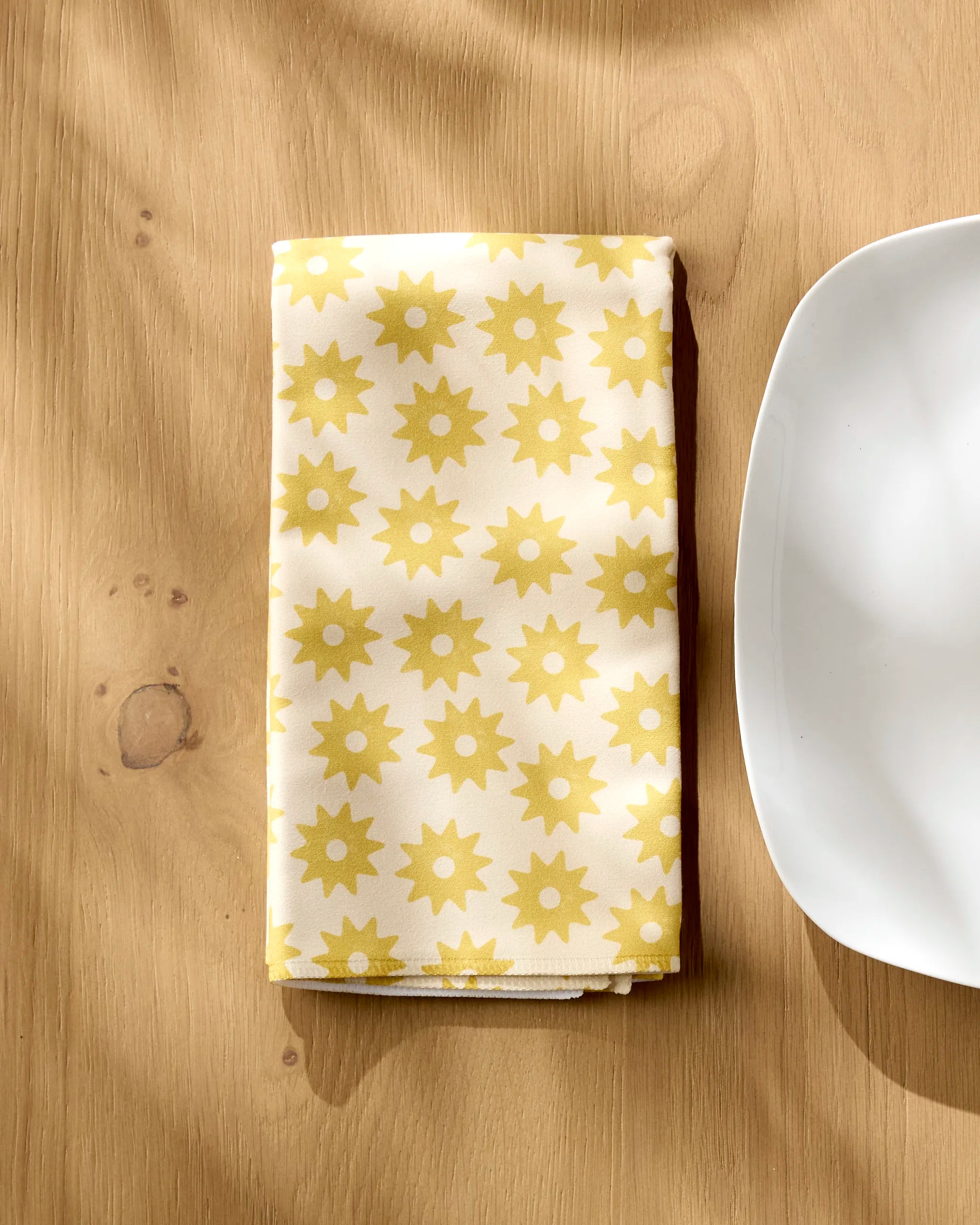 The Sunny Sun napkin by Geometry, featuring a yellow sunburst pattern, rests folded on a wooden table beside a white plate.