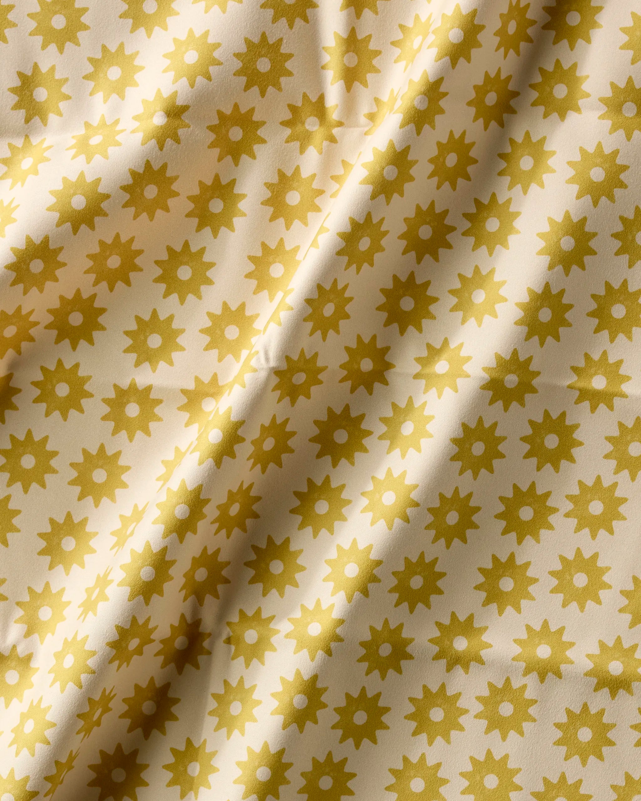 A close-up of Geometry’s Sunny Sun napkin set shows cream-colored fabric patterned with yellow sunbursts. Slightly wrinkled, this eco-friendly set brings soft folds—ideal for sustainable dining.