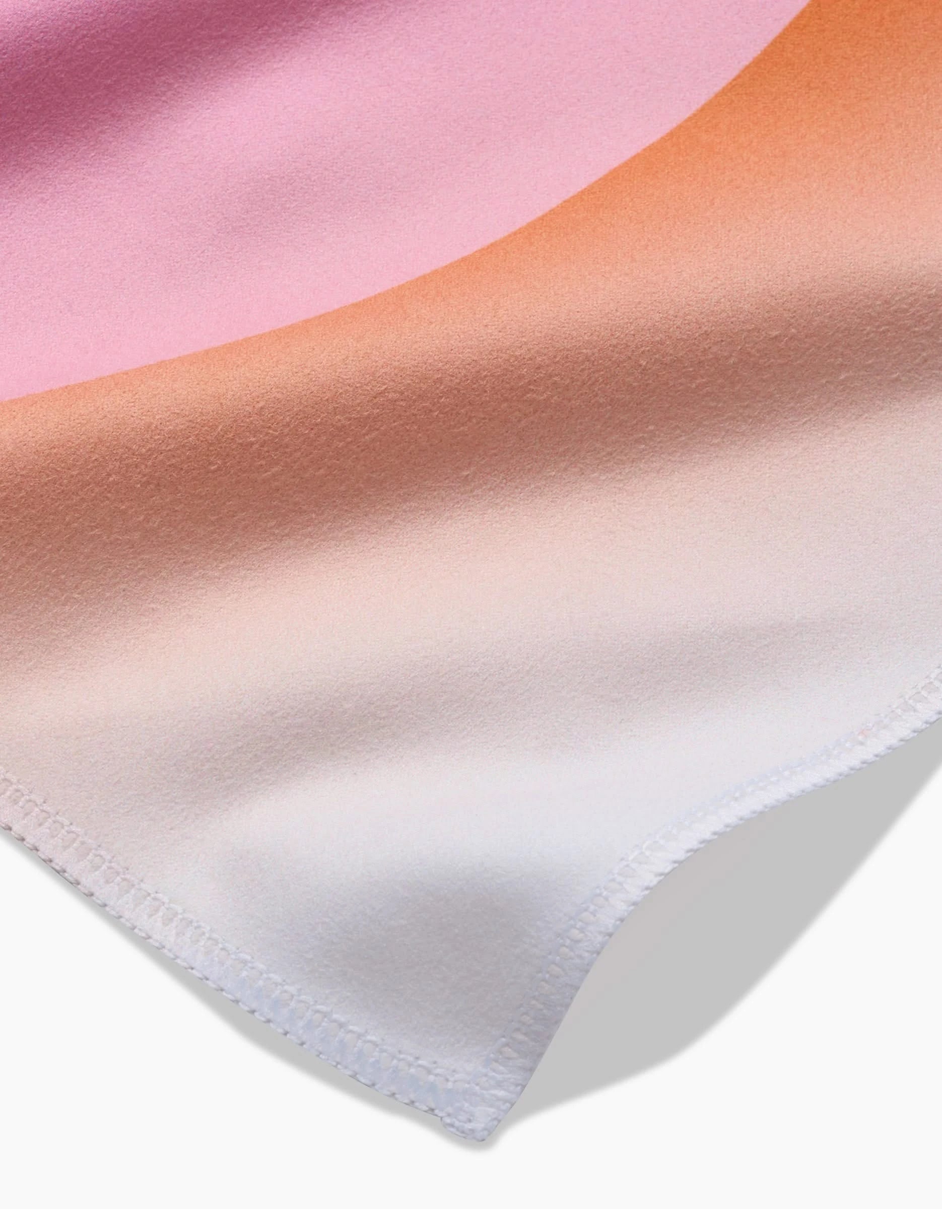 A close-up of GEOMETRY’s Sunrise napkins displays their absorbent fabric, gradient colors from pink to peach to white, and a neatly stitched edge casting a shadow on a white surface.