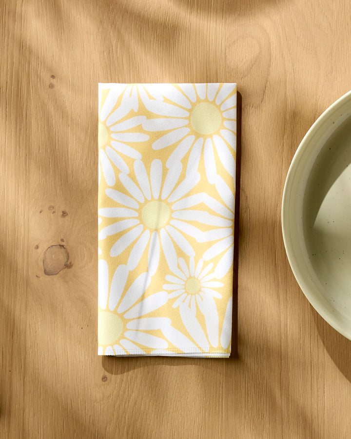 The Rebecca Bobko Sunshine Meadow reusable napkin, featuring a yellow and white daisy pattern, is folded on a light wooden table beside a cream-colored plate.