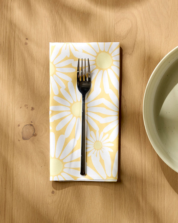 A fork rests on a folded Rebecca Bobko "Sunshine Meadow" reusable napkin with a yellow and white daisy pattern, placed on a light wooden table beside a cream-colored plate.