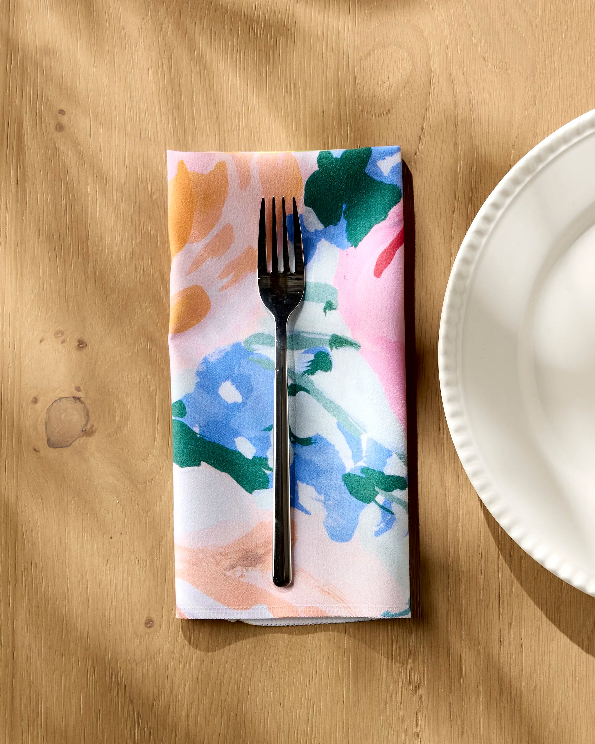 The Suzzy reusable napkin by Ania Zwara Art, featuring a colorful abstract pattern, sits beside a white scalloped-edge plate with a black fork on a light wooden table.