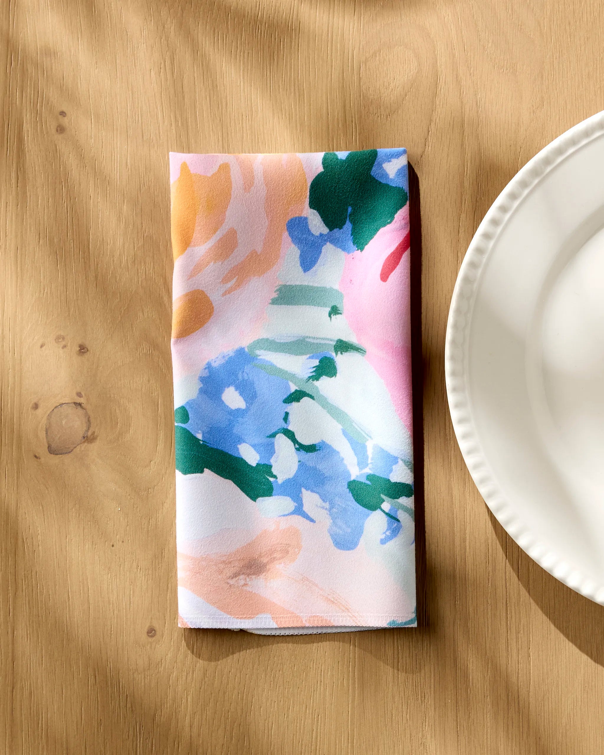 The Suzzy reusable napkin by Ania Zwara Art, featuring abstract multicolored patterns, is folded on a light wooden table beside a white scalloped plate.