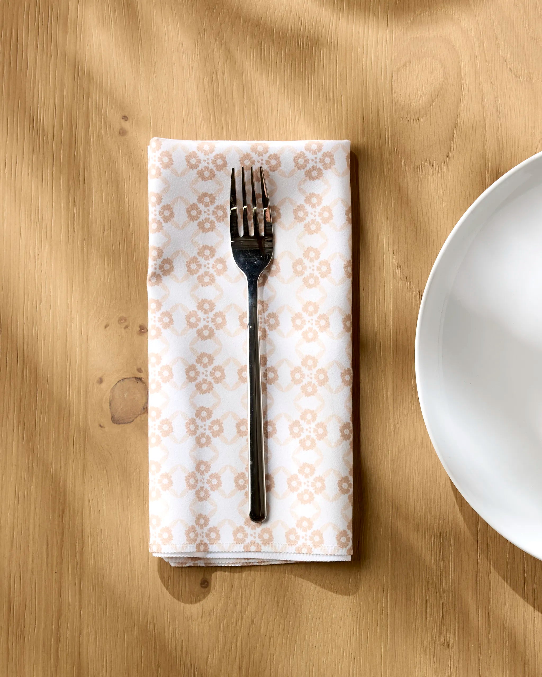 A Sweet Boho reusable table napkin by Hufton Studio is neatly folded on a wooden table beside a white plate, with a metal fork resting on top as sunlight casts soft shadows over the scene.