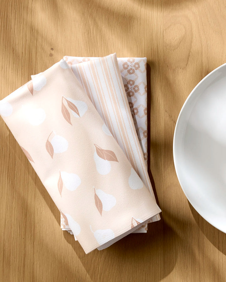 Three neatly folded Sweet Boho reusable napkins by Hufton Studio—one with pears, one striped, and one floral—are placed on a light wooden table beside a white plate in soft natural light.